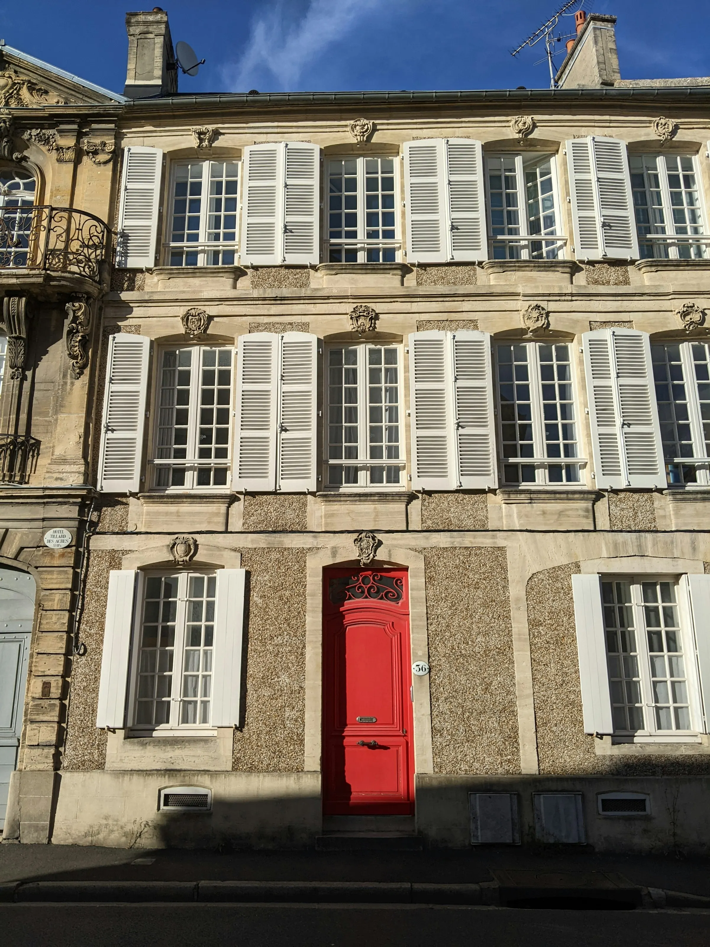 A handsome 18th-century Bayeux townhouse with a carved limestone facade, rows of white louvered shutters and a vivid red front door, warm low sun raking across the stone.