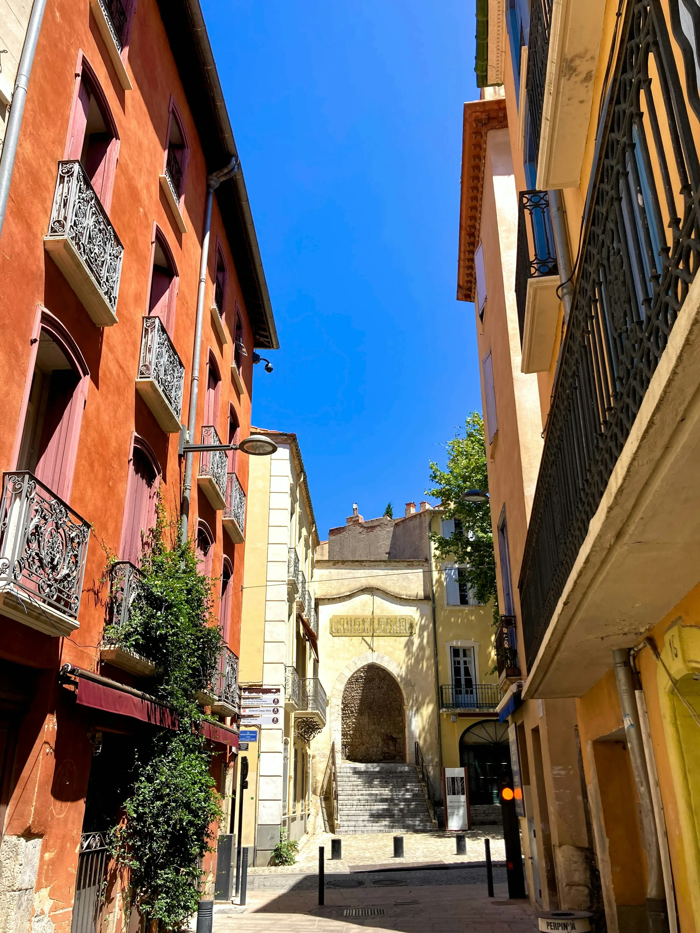 A sunlit narrow lane in Perpignan's old town lined with warm ochre and terracotta houses with wrought-iron balconies, leading to a stone archway and steps under a deep blue sky.