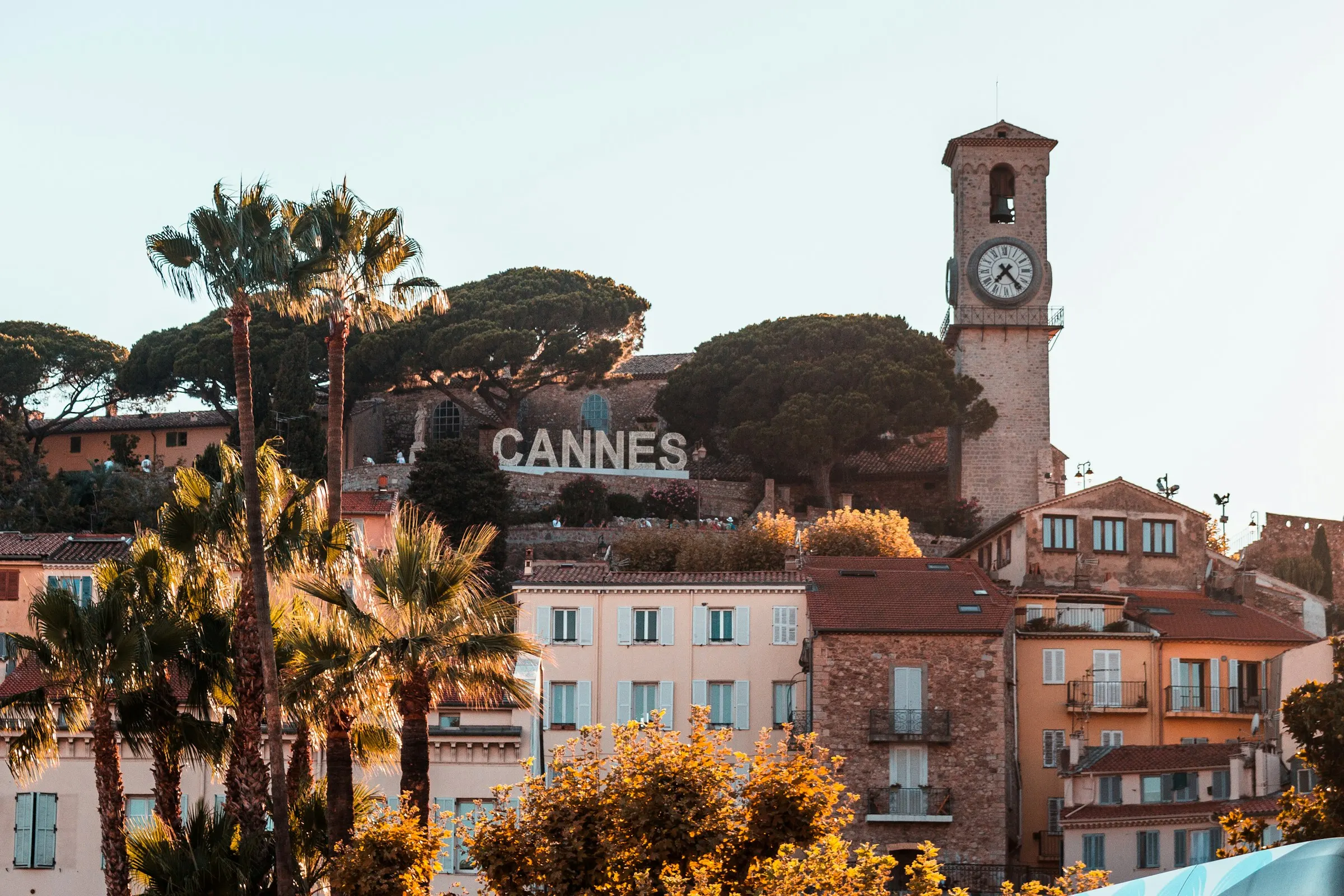 Golden evening light on Le Suquet, the old quarter of Cannes — the stone Notre-Dame-d'Esperance clocktower and the hillside CANNES sign above ochre houses, umbrella pines and swaying palm trees.
