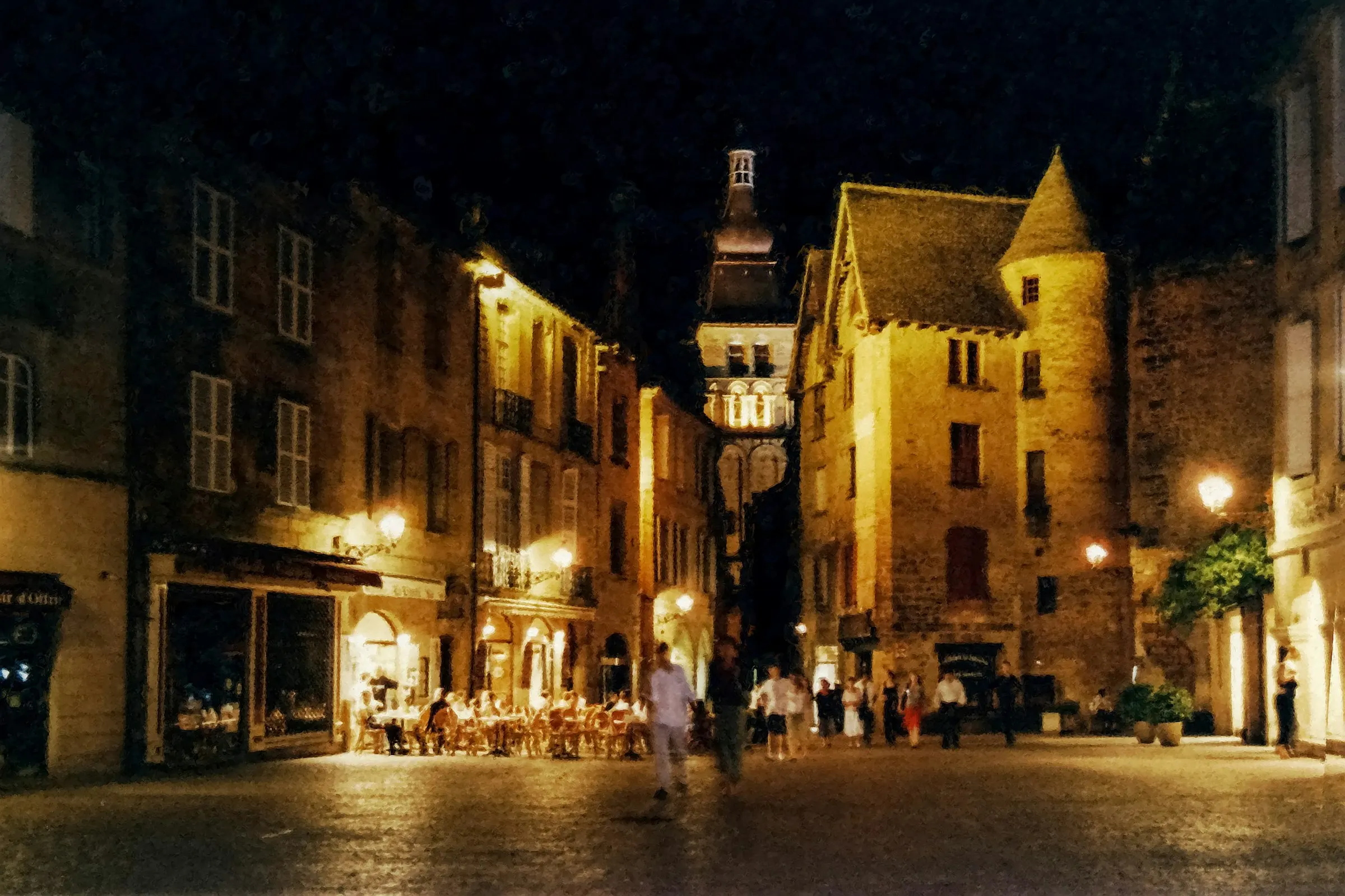 A warm evening on a Sarlat square, cafe tables and diners glowing under lamplight beneath a turreted honey-stone mansion as people stroll the cobblestones