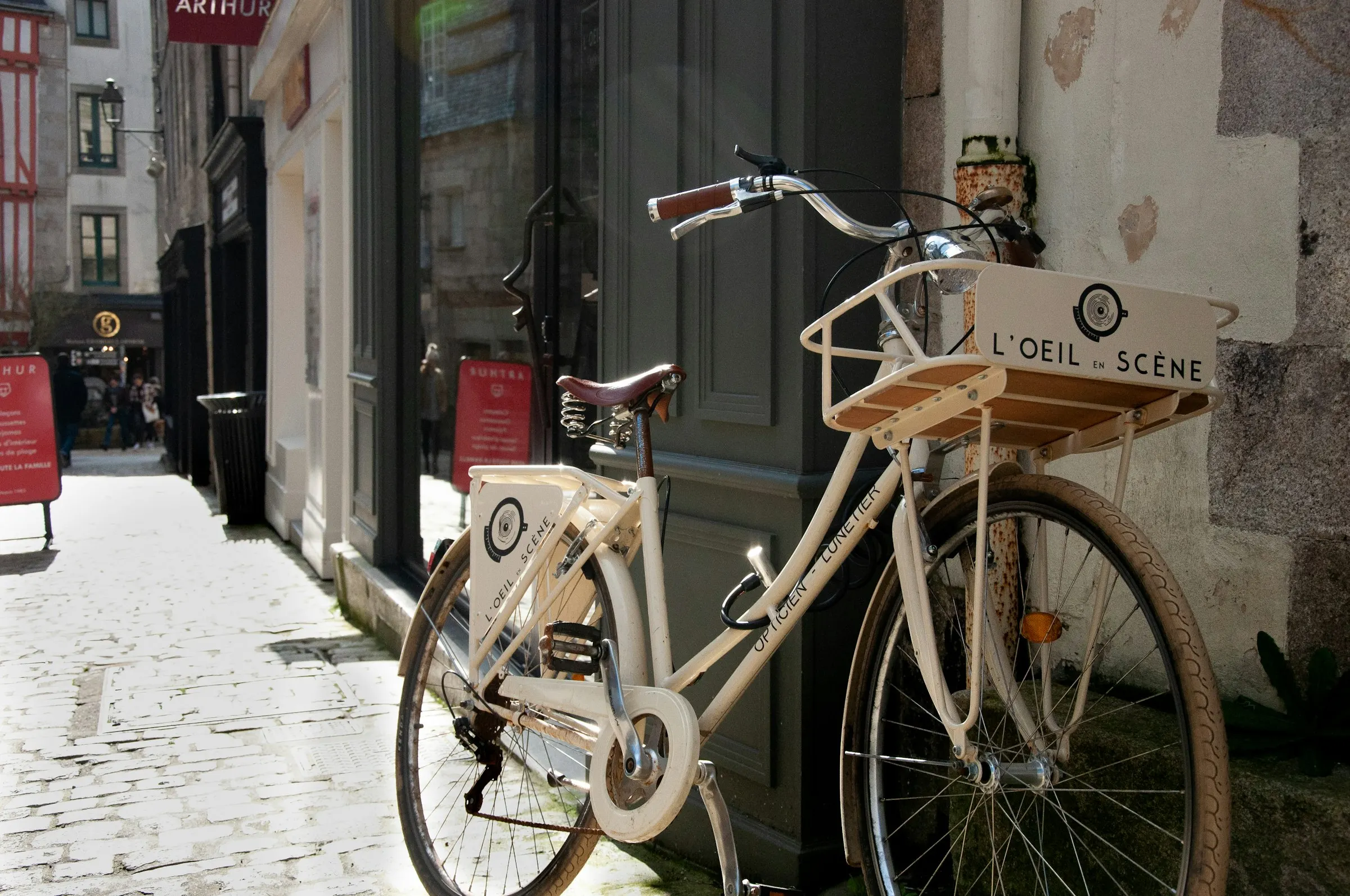 A vintage cream bicycle with a wooden front rack leans against a granite corner on a sunlit, cobbled Quimper lane, half-timbered facades receding into the background.