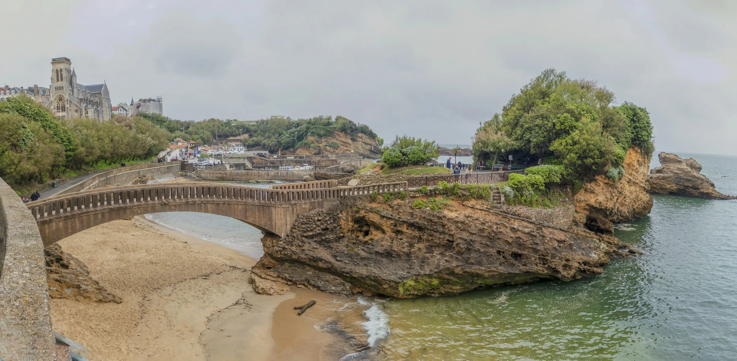 Biarritz seafront panorama with the arched stone footbridge crossing to the Rocher du Basta sea-rock, the Eglise Sainte-Eugenie and old-town rooftops on the hill above a small sandy cove