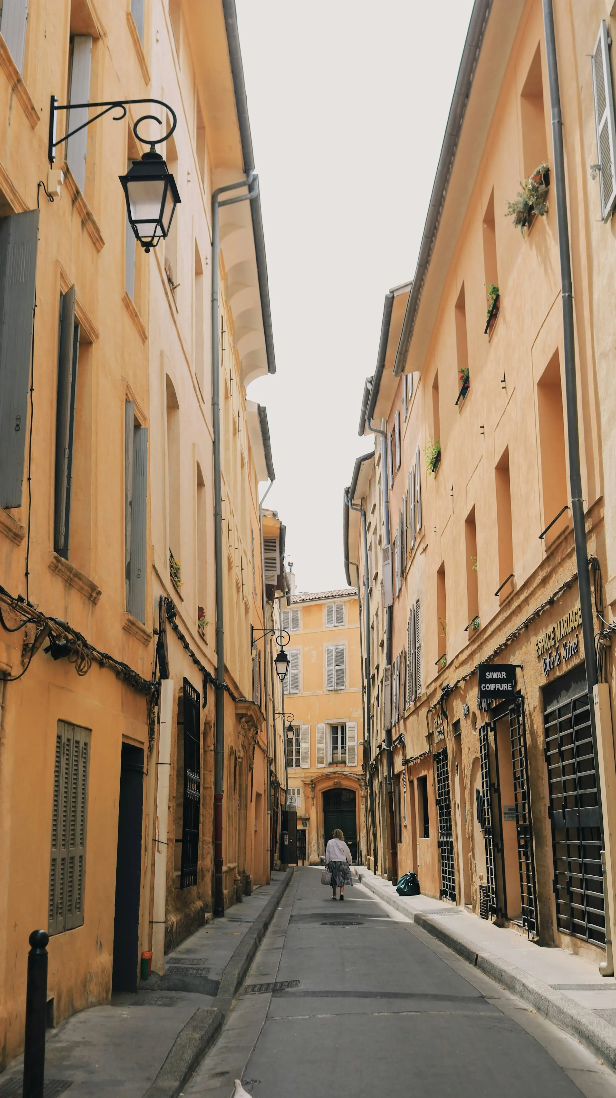 A narrow, sunlit old-town lane in Aix-en-Provence lined with ochre shuttered buildings and black wrought-iron lanterns, a lone shopper walking the quiet street.