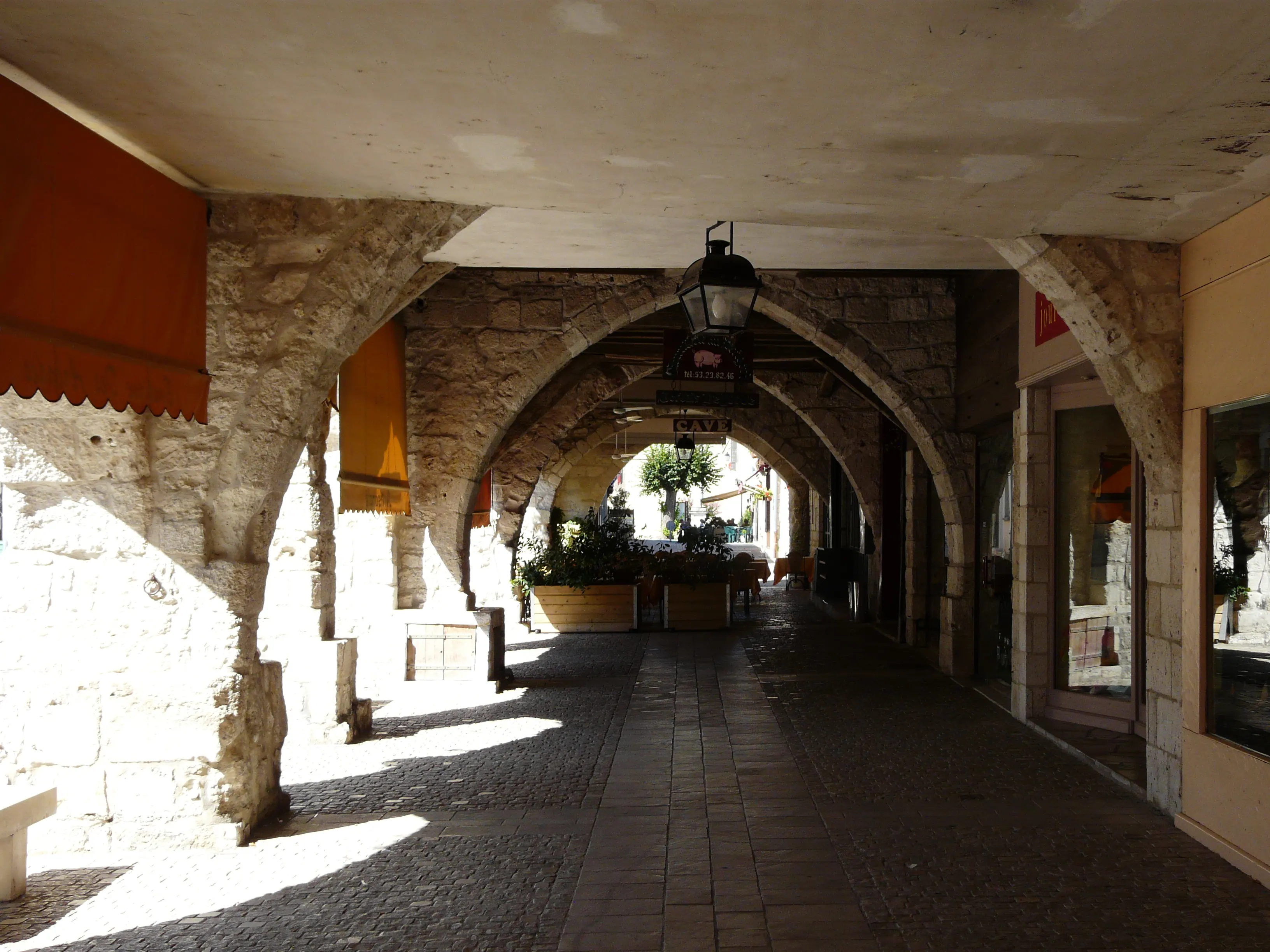 Sunlight falling through the medieval stone arcades (couverts) that ring Eymet's bastide square, past a restaurant terrace and a wrought-iron lantern