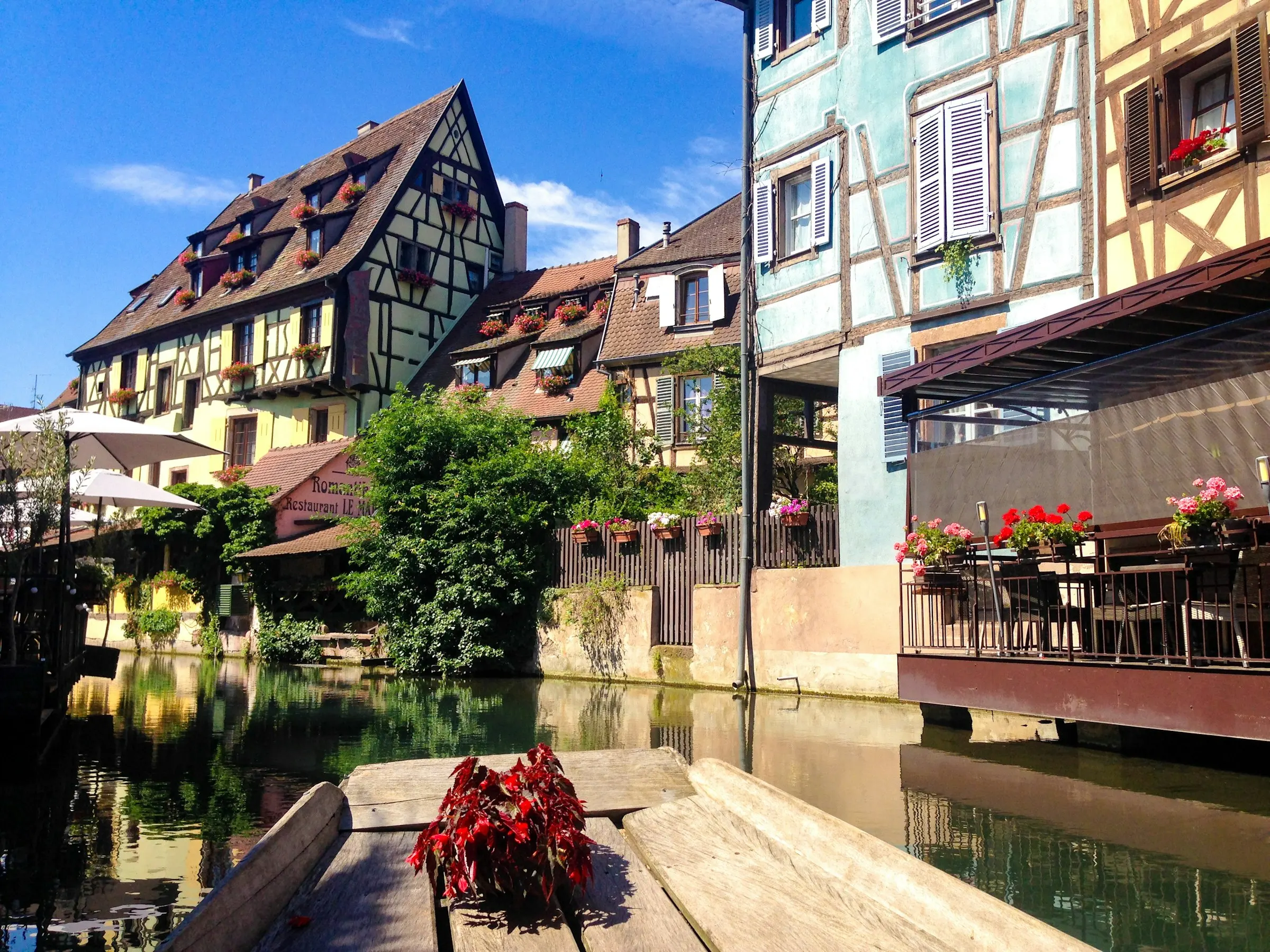 View from a flat-bottomed boat drifting along a Petite Venise canal in Colmar, cafe terraces and timbered houses on the banks