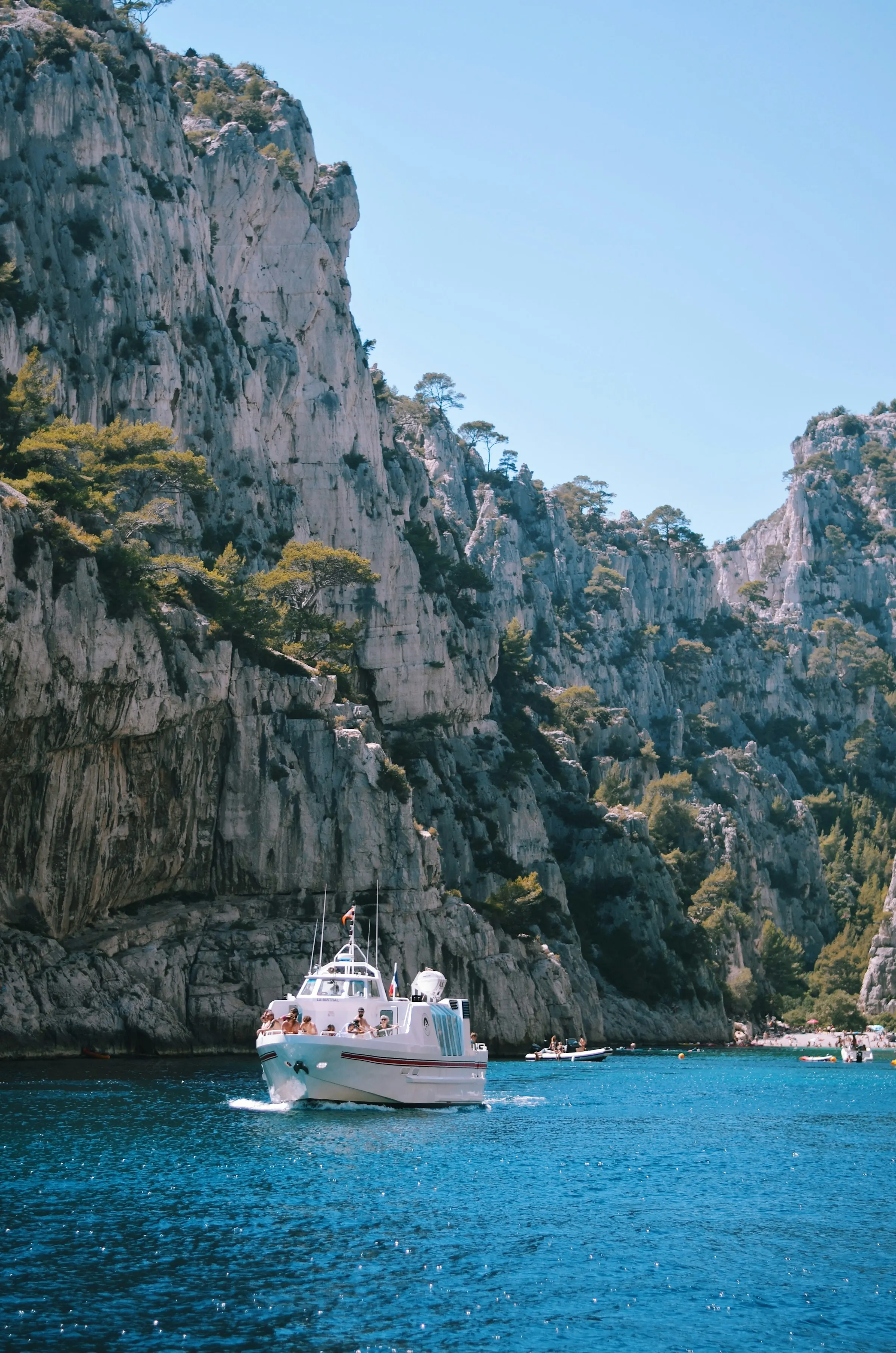 A small white tour boat cruising the deep-turquoise water of a Calanque inlet near Marseille, dwarfed by towering pale limestone cliffs dotted with pines under a clear summer sky.