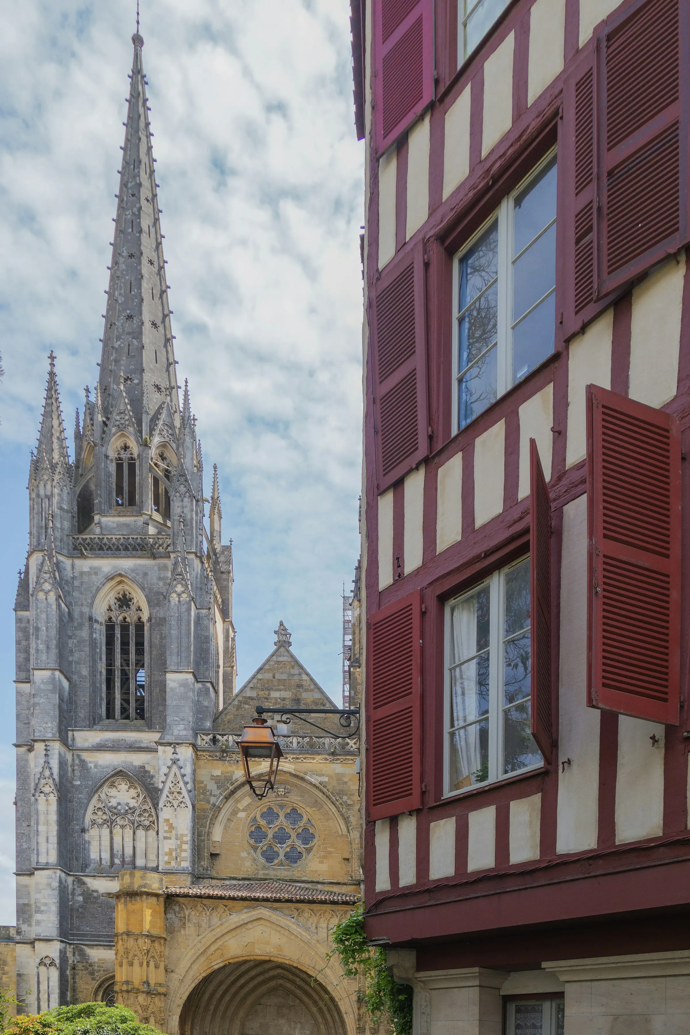 The soaring Gothic stone spire of Bayonne's Cathedrale Sainte-Marie rising under a blue sky beside the red shutters and cream timber framing of a half-timbered house.