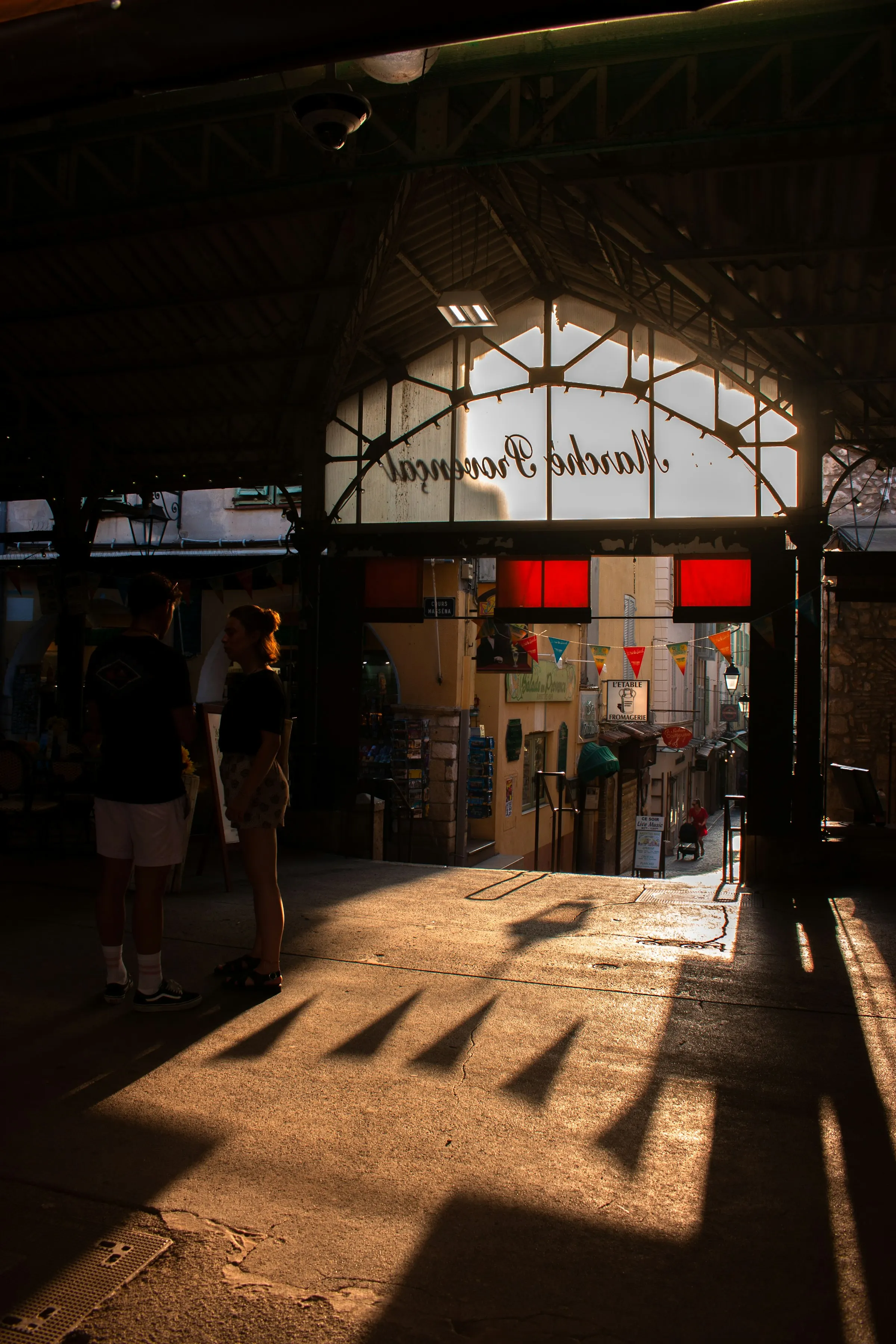 Golden late-day light streaming through the wrought-iron entrance of the covered Marche Provencal in Antibes' old town, two shoppers in silhouette, with a fromagerie and Provencal shop signs down the lane beyond.