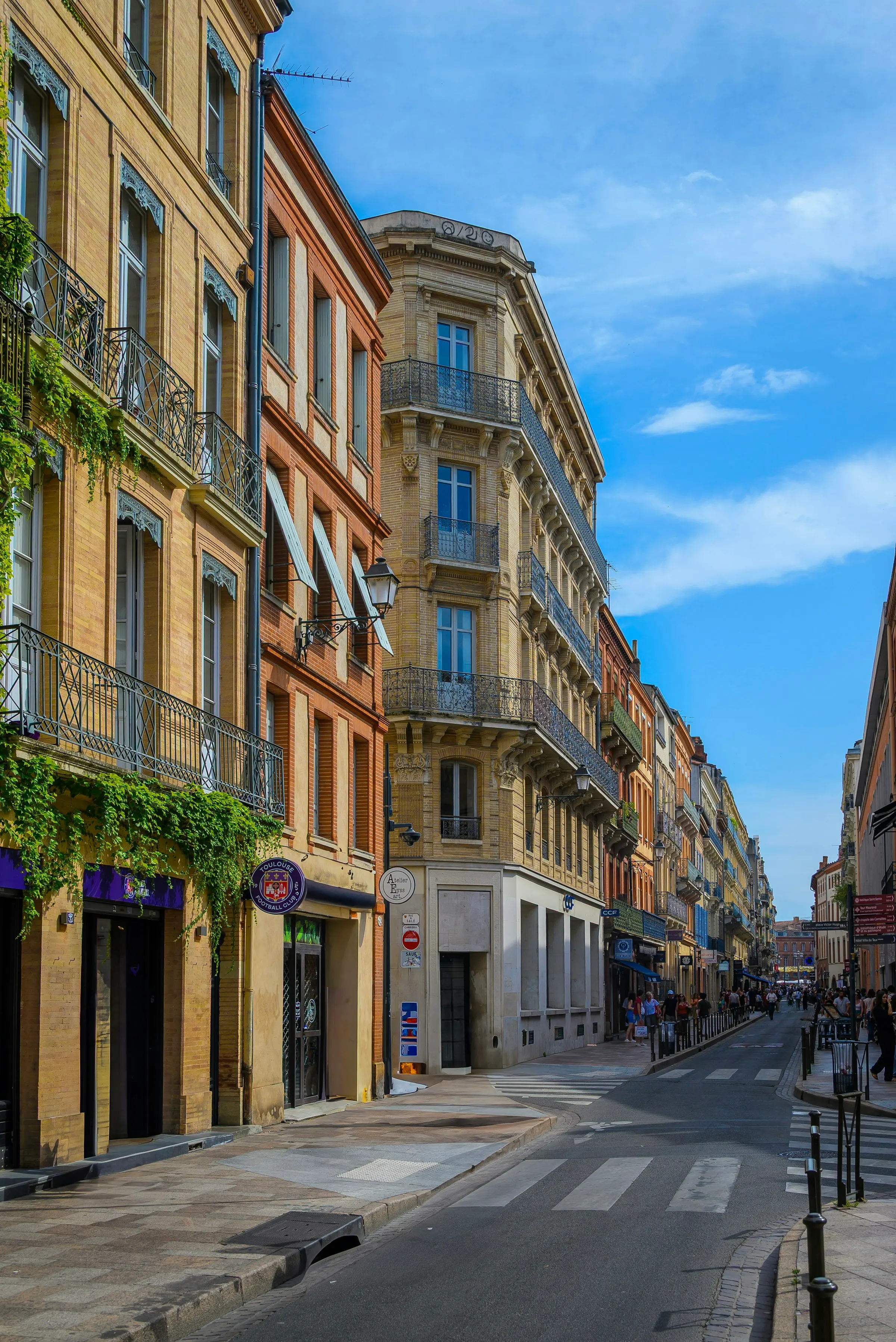 A sunlit central Toulouse street lined with pink-brick and stone townhouses with wrought-iron balconies and trailing greenery, a Toulouse Football Club shopfront and pedestrians strolling toward the old town.