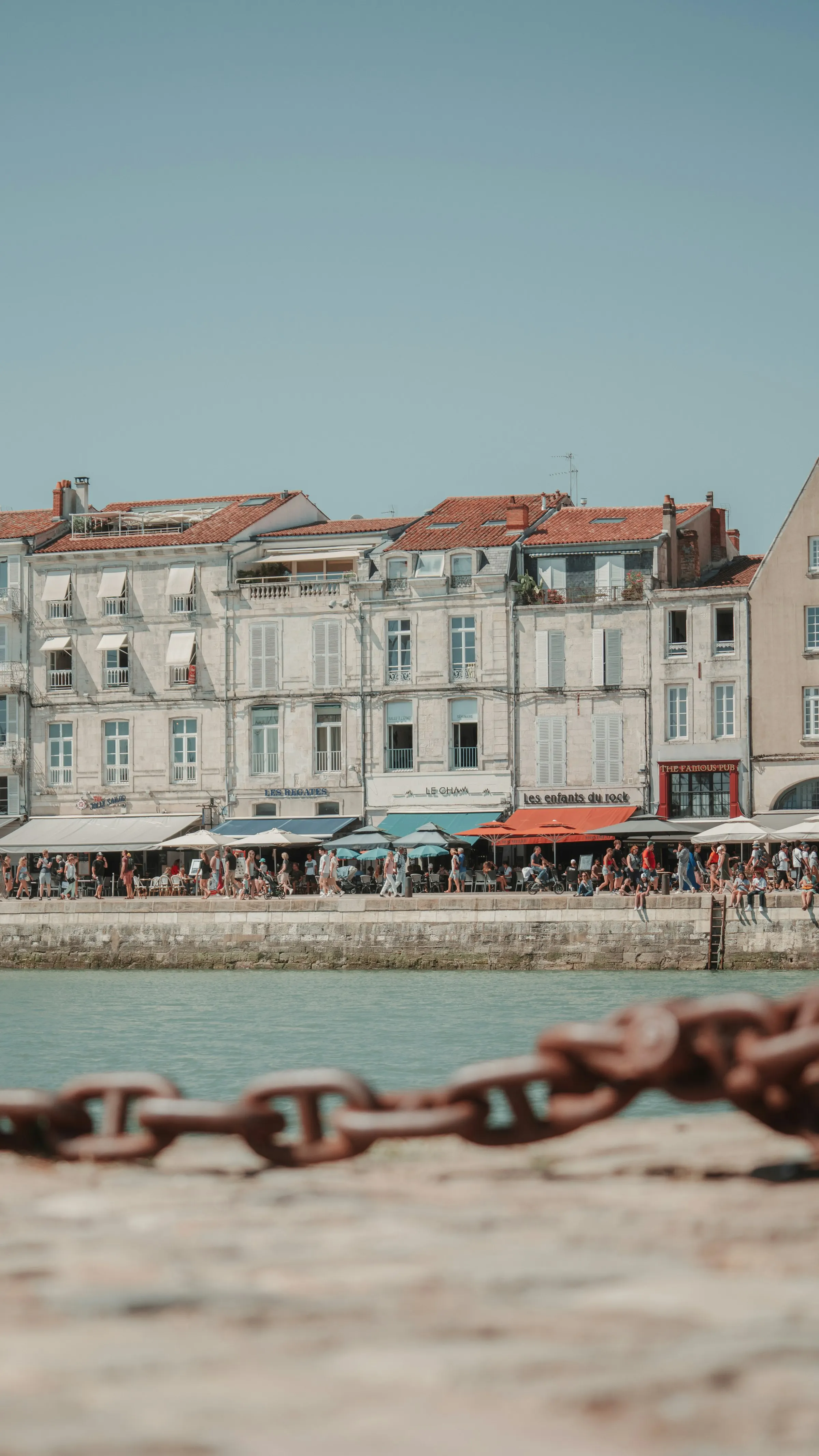Sunlit quay of La Rochelle's old port lined with busy café and restaurant terraces under white parasols, a rusted iron mooring chain strung across the warm stone dock in the foreground.
