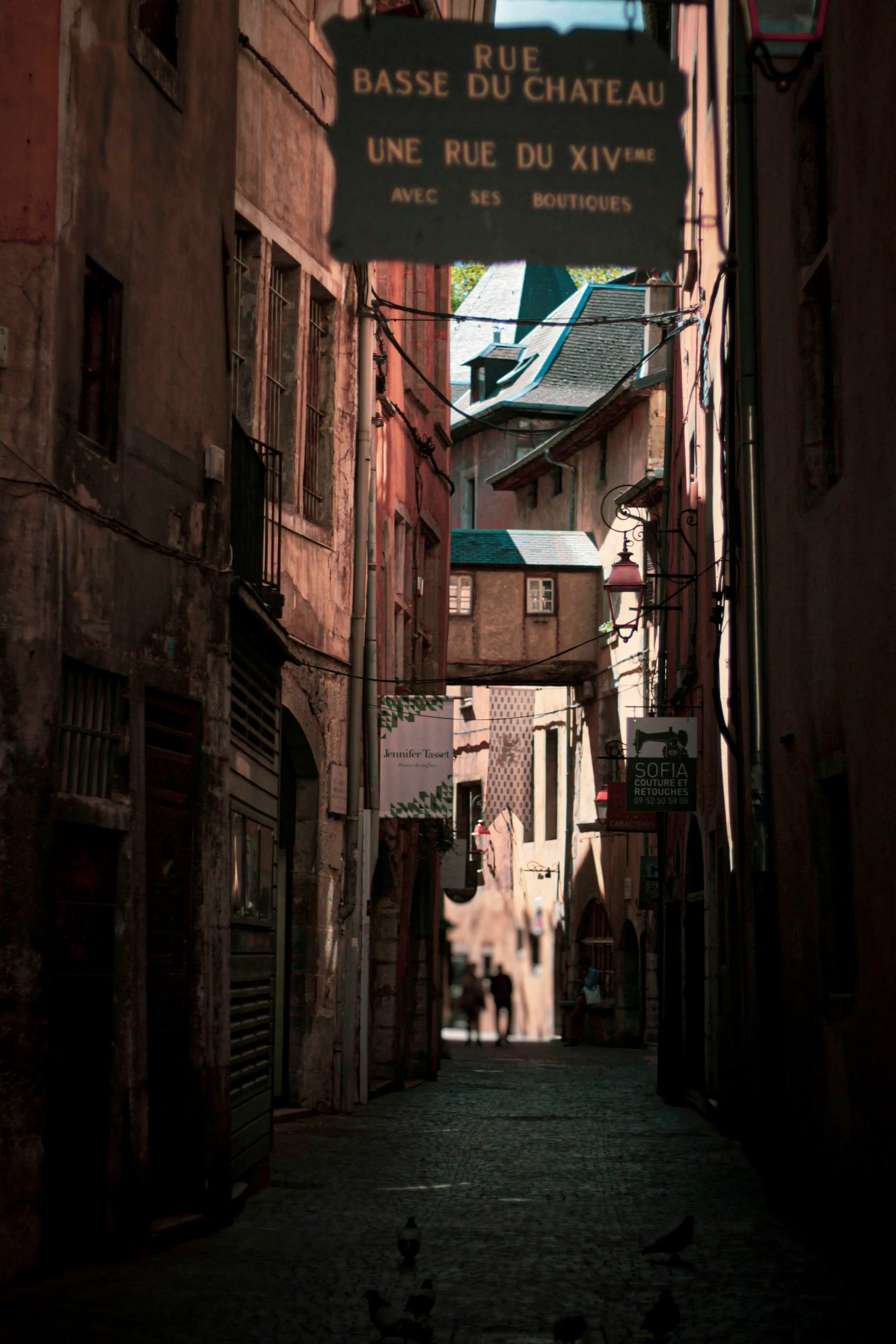 A narrow cobbled lane in Chambery's medieval old town beneath a hanging sign reading 'Rue Basse du Château, une rue du XIVeme avec ses boutiques', tall shuttered stone houses leaning in and a few strollers in the sunlit distance.