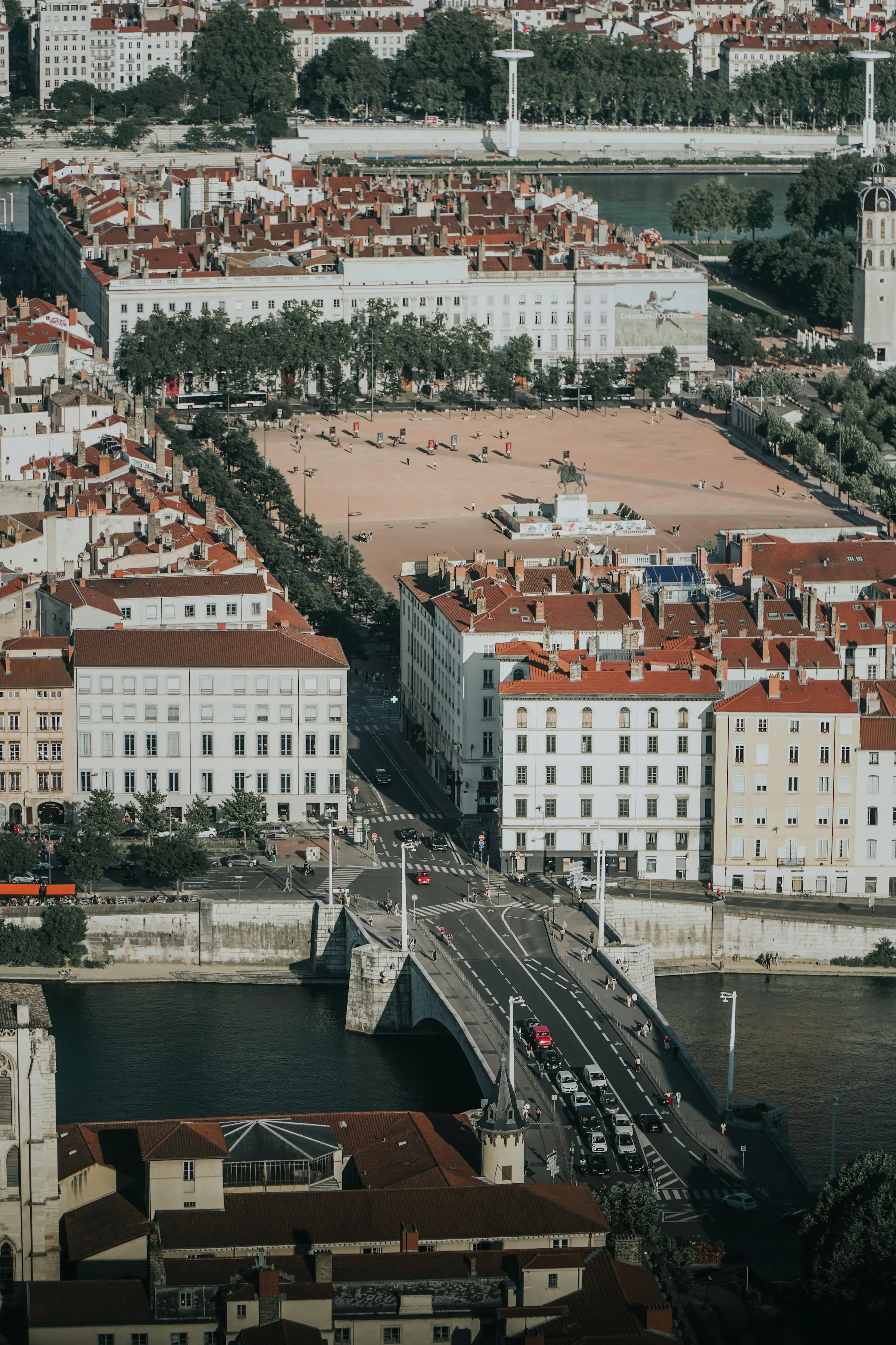 Aerial view of the vast rust-red expanse of Place Bellecour with the Louis XIV equestrian statue at its center, framed by a stone bridge over the Rhone and rows of red-roofed Lyon buildings