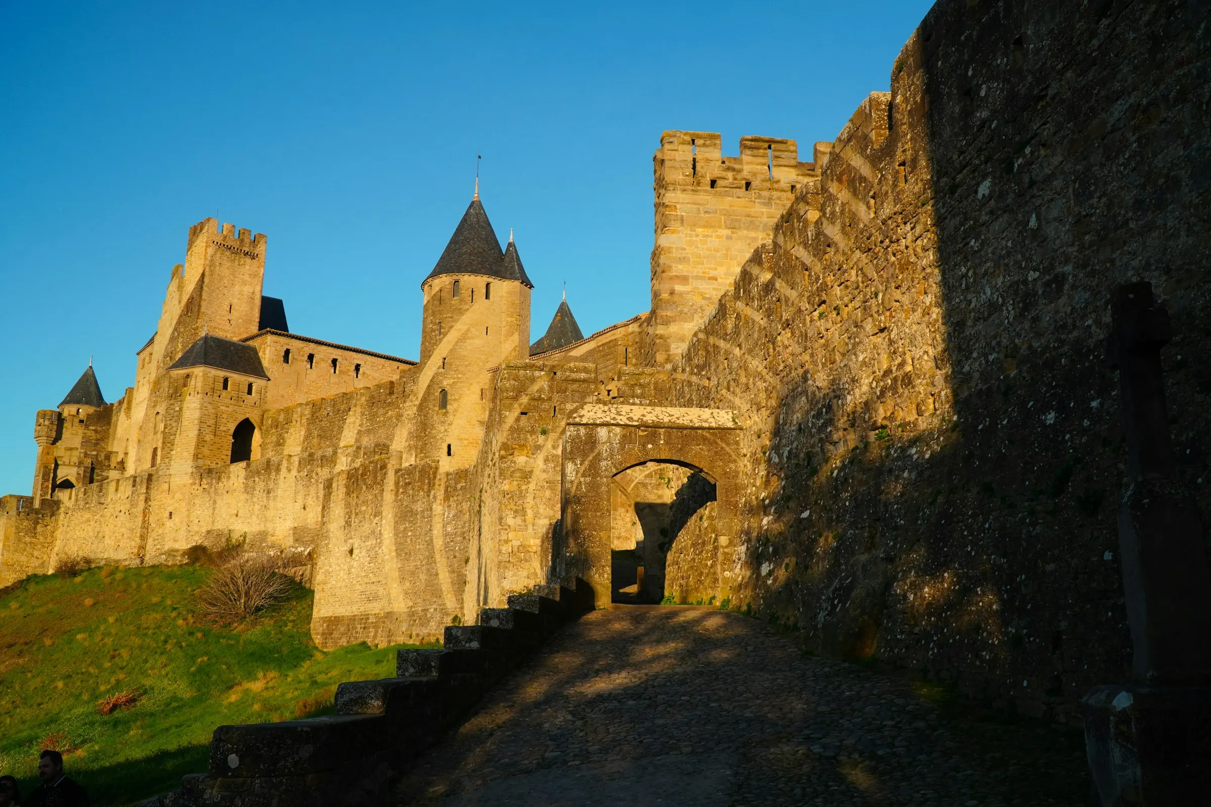 Warm evening light on the cobbled ramp climbing to a fortified stone gateway of Carcassonne, crenellated ramparts and towers above