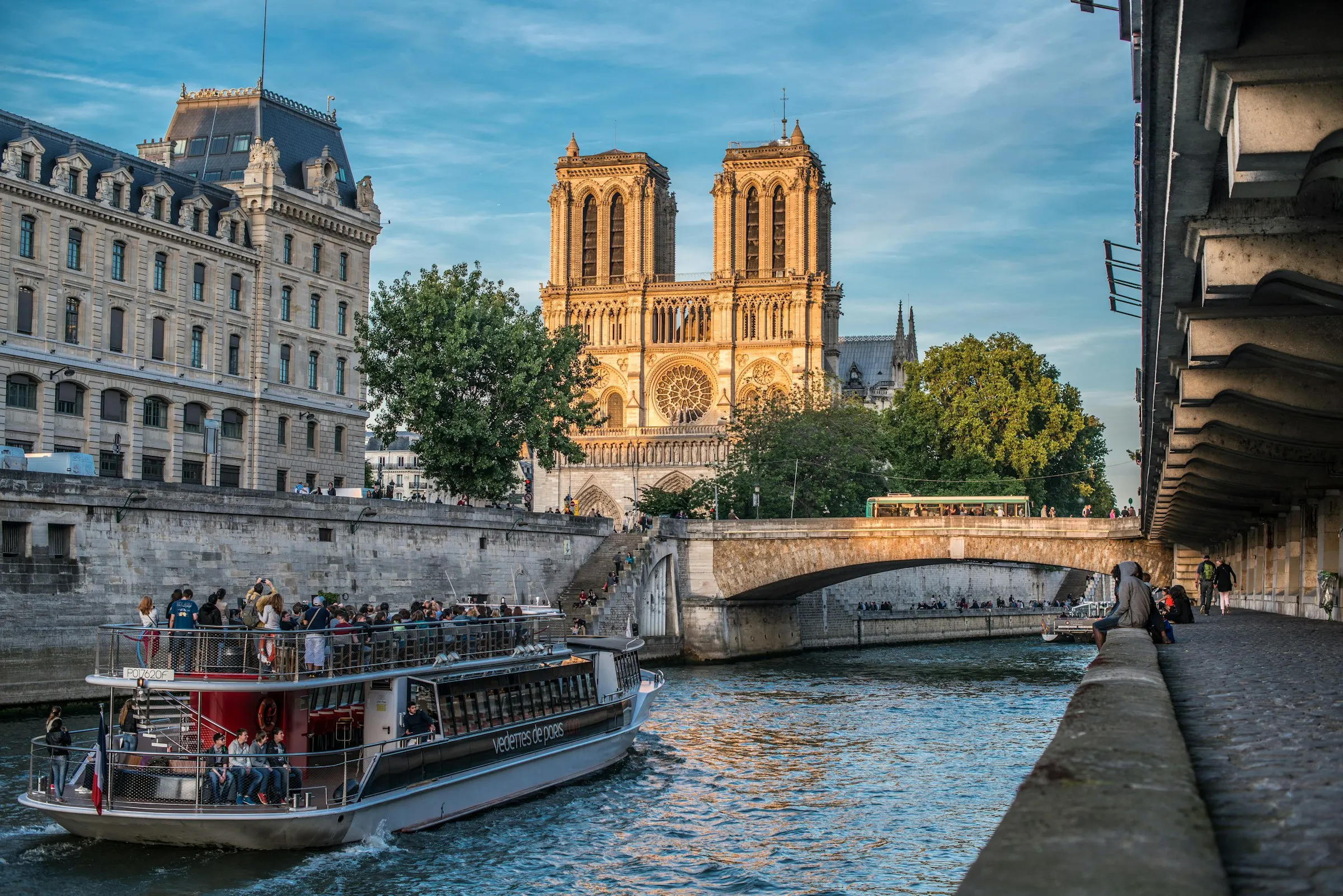 Notre-Dame de Paris glowing gold at sunset above the Seine as a Vedettes de Paris sightseeing boat passes beneath a bridge