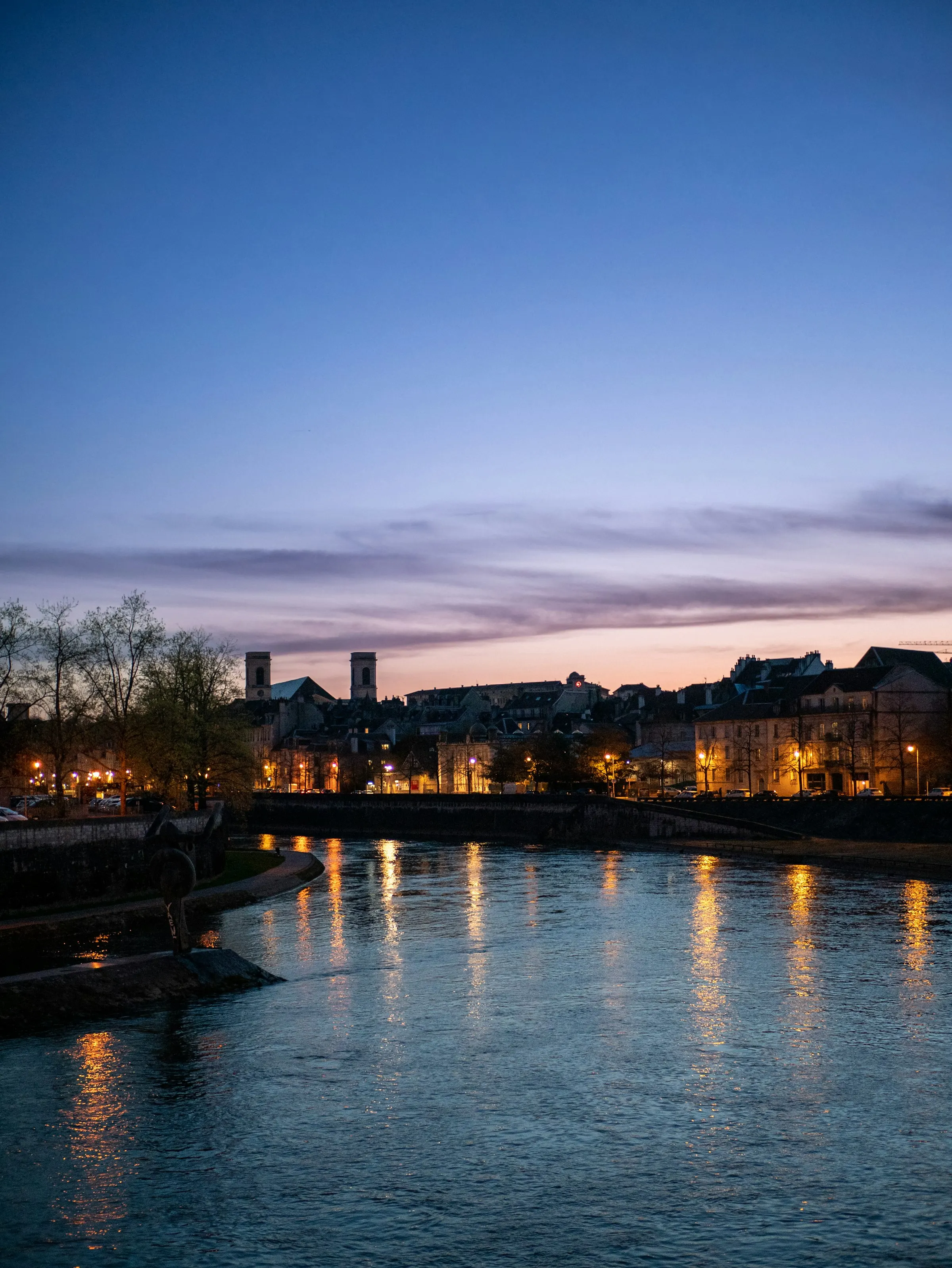 The Doubs river at blue hour, its calm surface catching long gold reflections of quayside lamps below Besançon's old-town skyline and the twin stone towers of the Madeleine church against a violet sky.