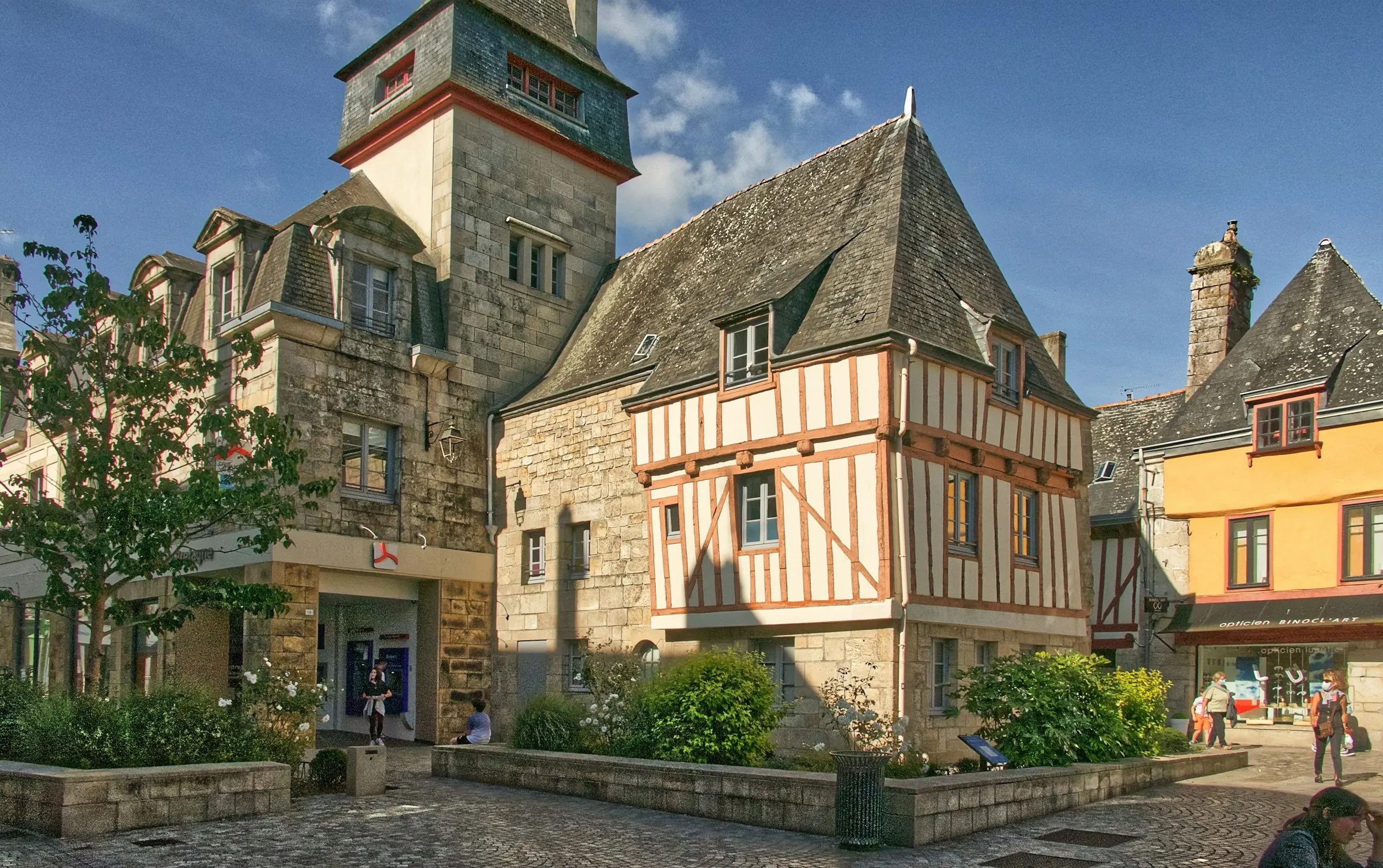 A sunlit cobbled square in Quimper's old town with a granite belfry tower and a red-timbered, slate-roofed half-timbered house, planted beds and a few strollers in warm afternoon light.