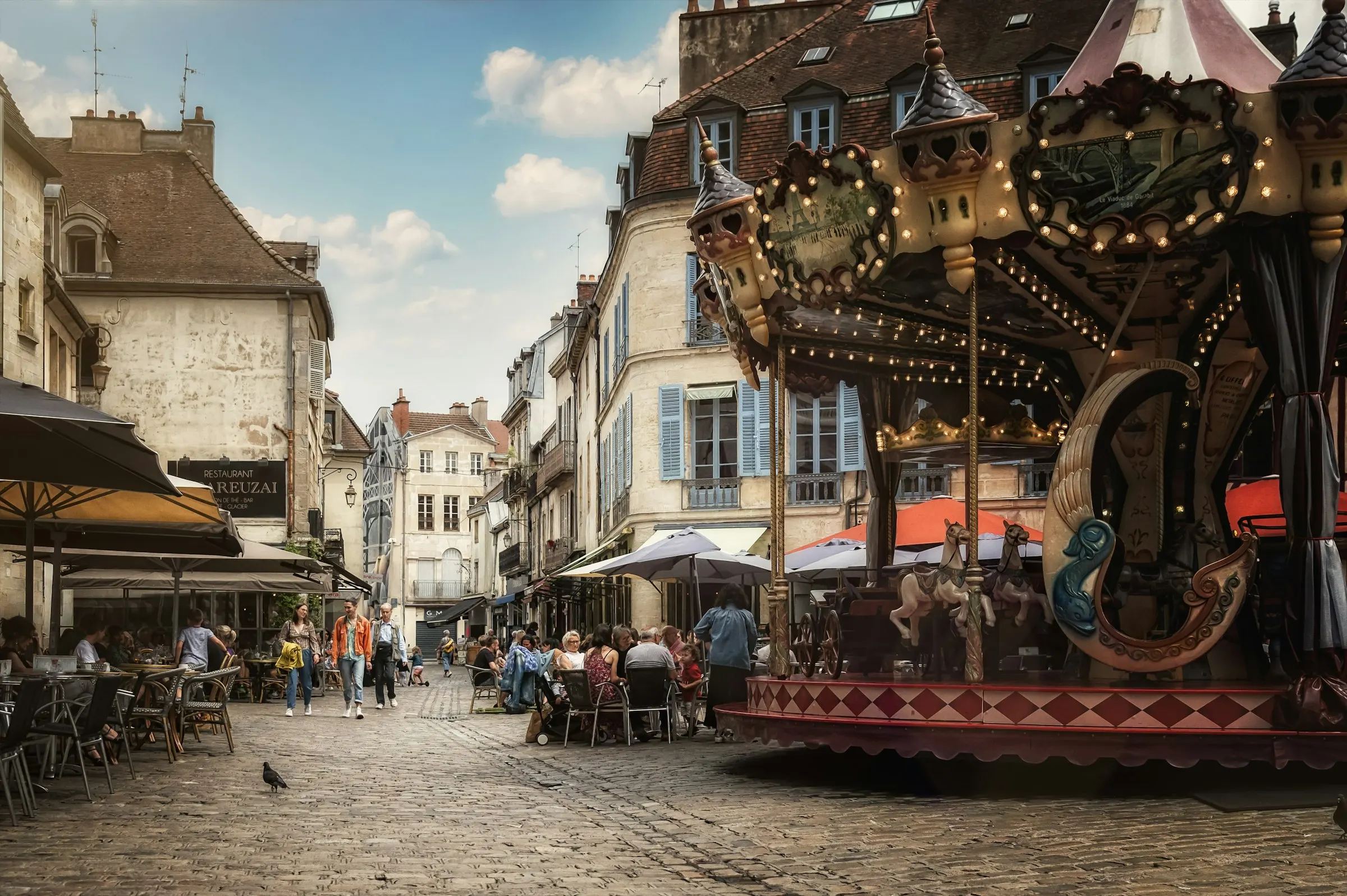 A vintage carousel and busy cafe terraces on Dijon's cobbled Place Francois Rude, people dining among stone and half-timbered Burgundian buildings