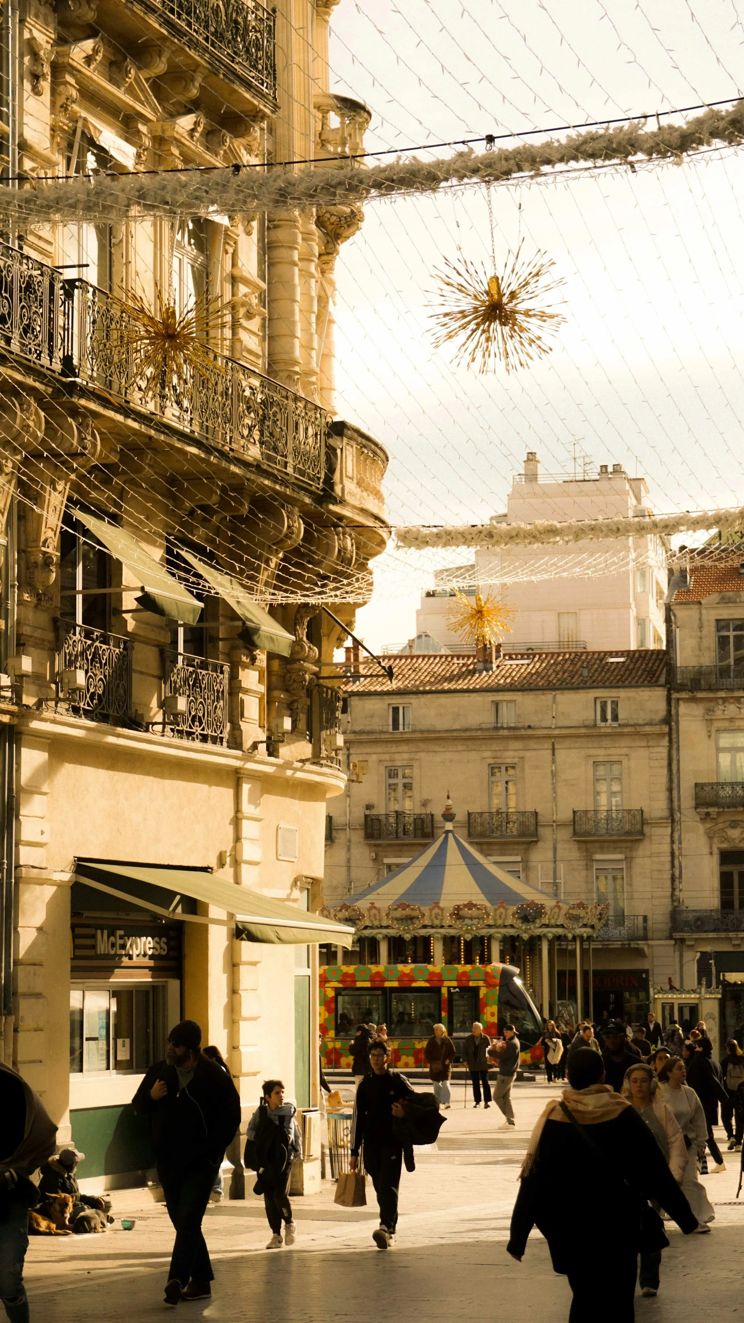 A brightly painted Montpellier tram passes a carousel and cafe terraces on a pedestrian street lit by golden late-afternoon sun, with shoppers strolling beneath festive lights.