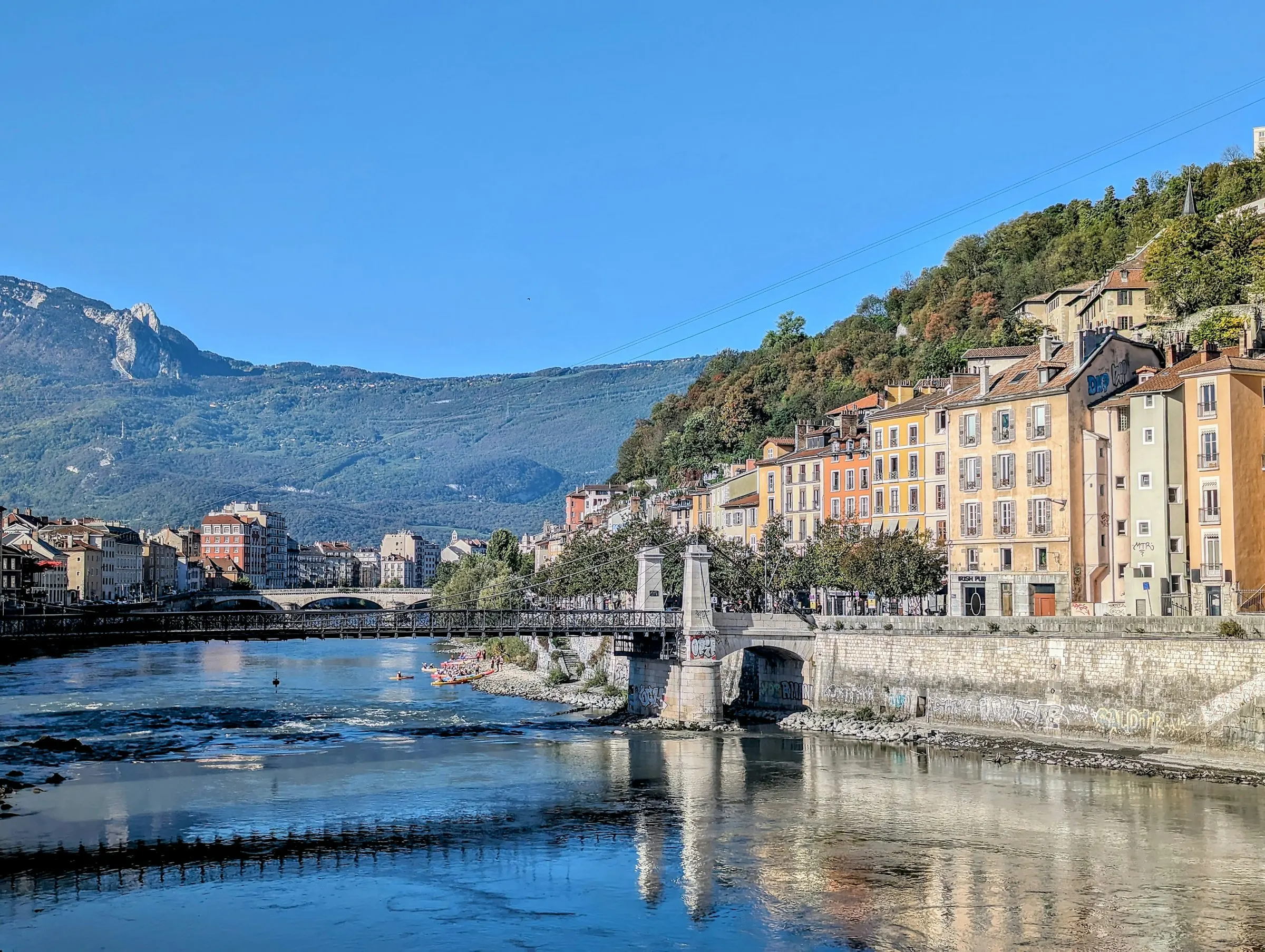 The Isere river quays in Grenoble's old town, lined with tall ochre and pastel façades, a slim white suspension footbridge and a stone arch bridge crossing the water, kayakers paddling below a forested mountain ridge.