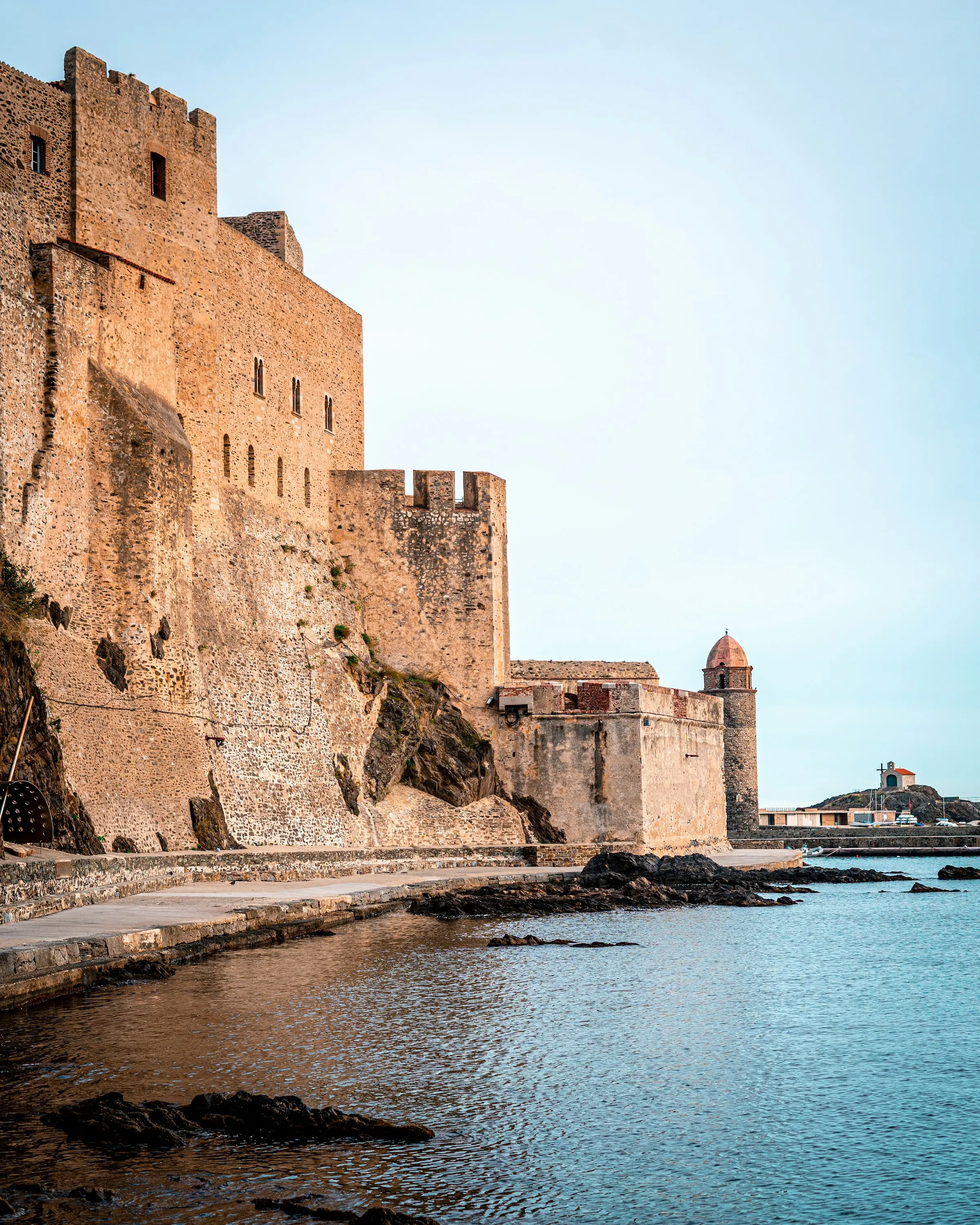 The massive sandstone ramparts of Collioure's Chateau Royal glowing at golden hour above the water's edge, the bell tower visible beyond