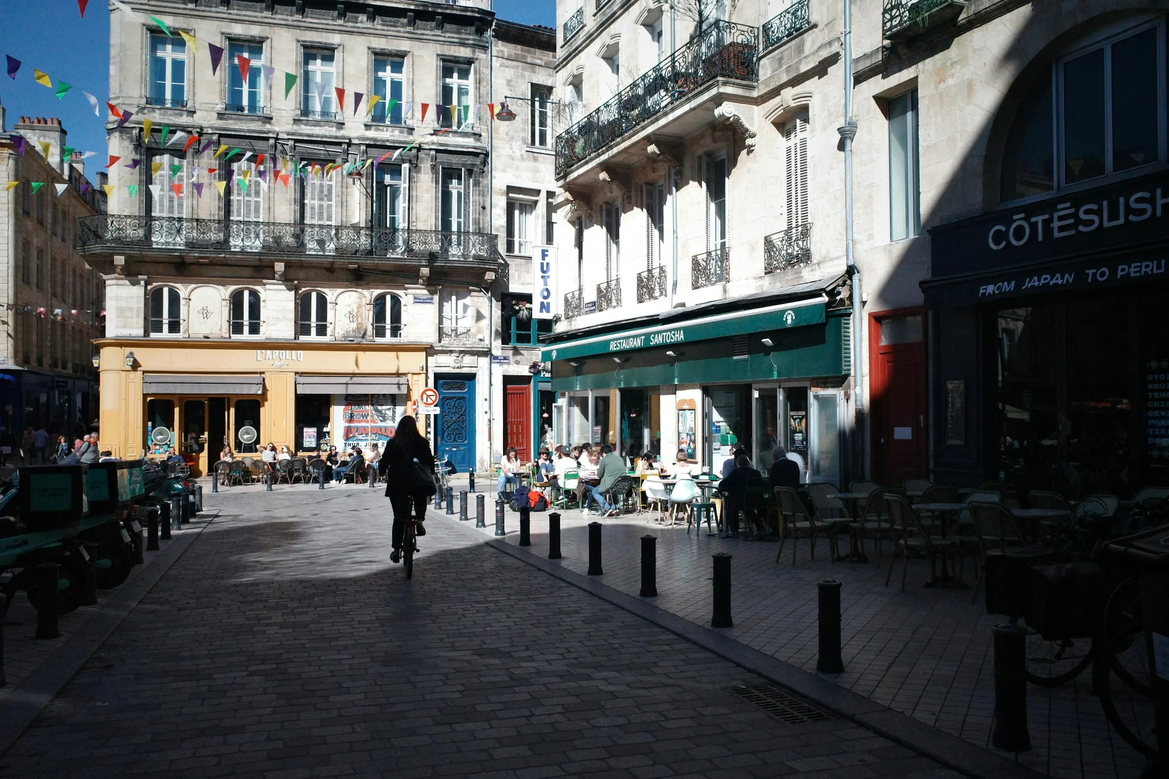 A sunlit pedestrian square in Bordeaux's old town, café terraces spilling onto pale-stone paving beneath strings of colorful bunting, a cyclist rolling past cream 18th-century facades with wrought-iron balconies.