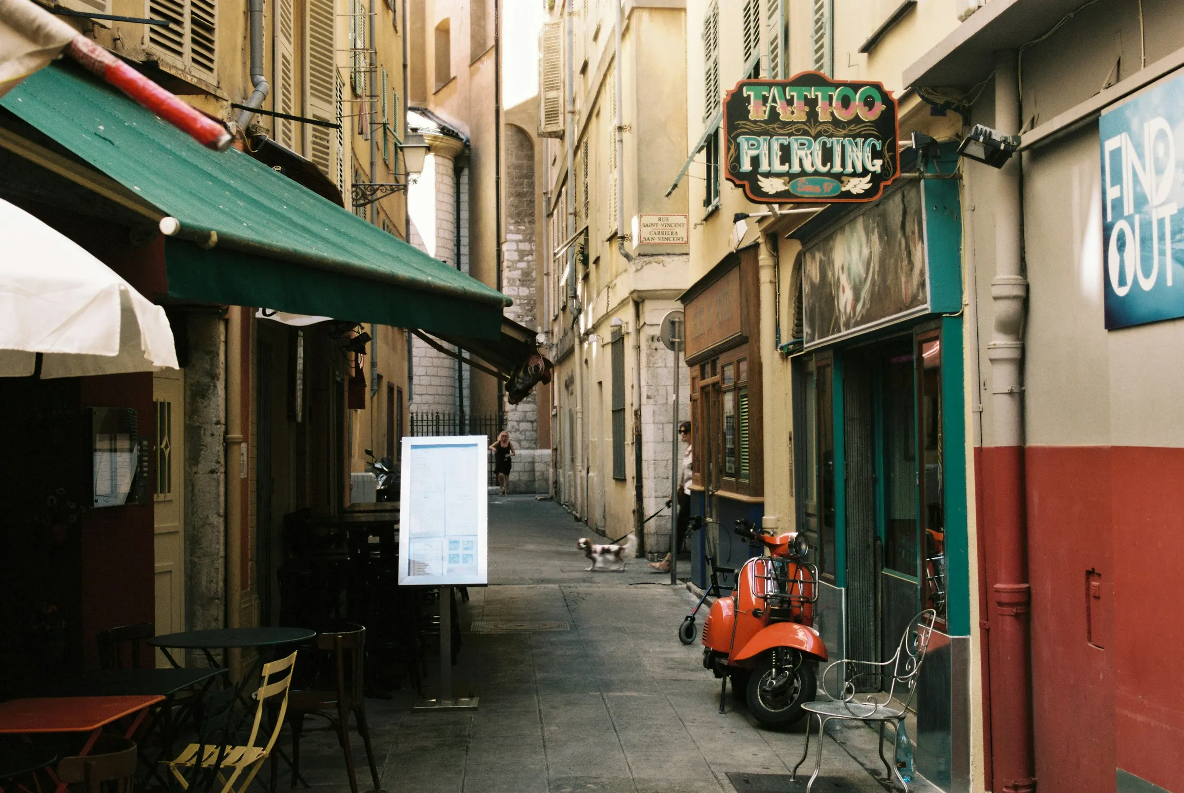 A narrow lane in Vieux Nice with cafe tables and a parked scooter, old-town facades in warm light