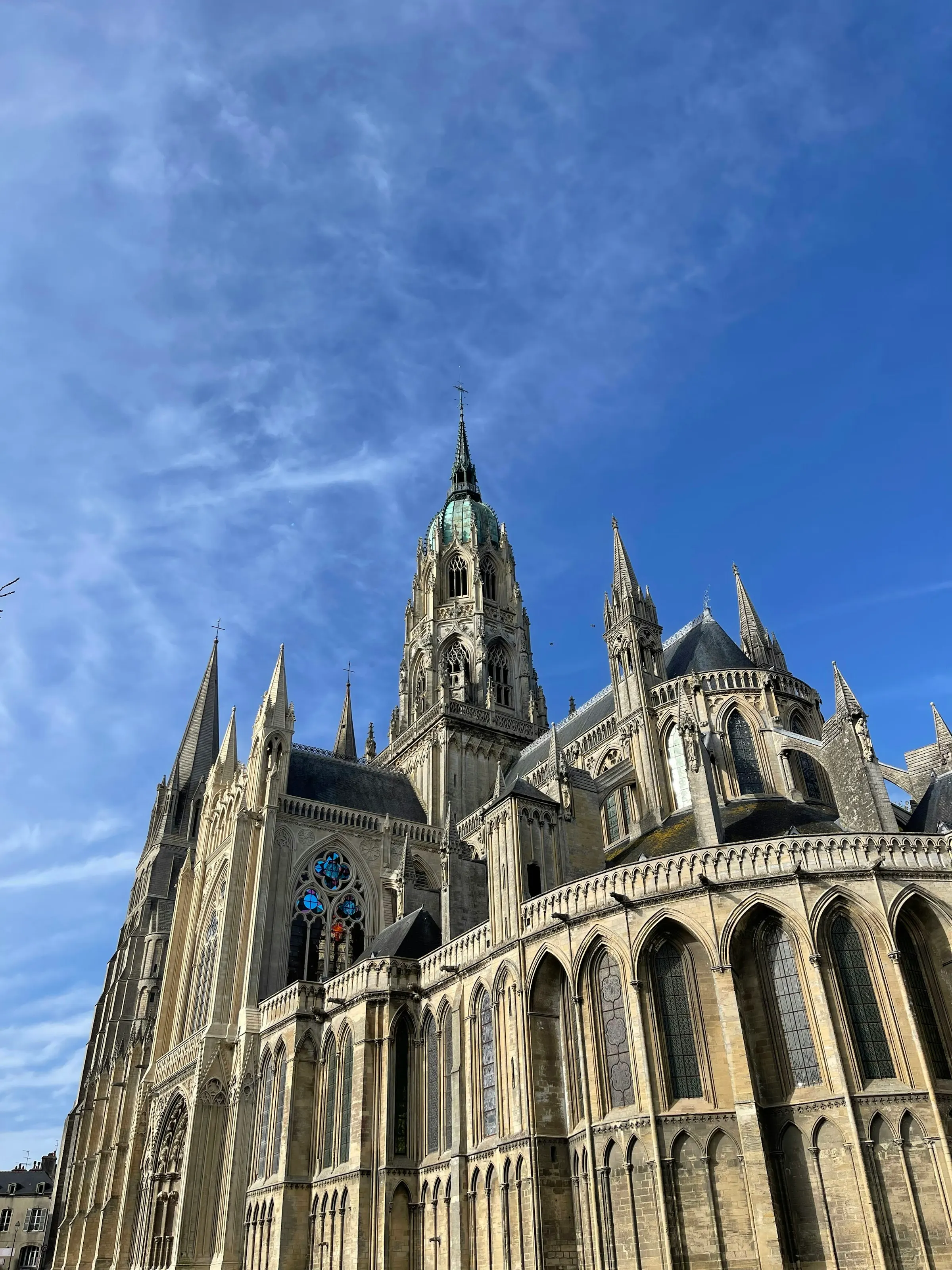 The Gothic Notre-Dame de Bayeux cathedral rising against a deep blue sky, its central tower crowned by a green copper dome and ringed by pale stone spires, pinnacles and arched flying buttresses.