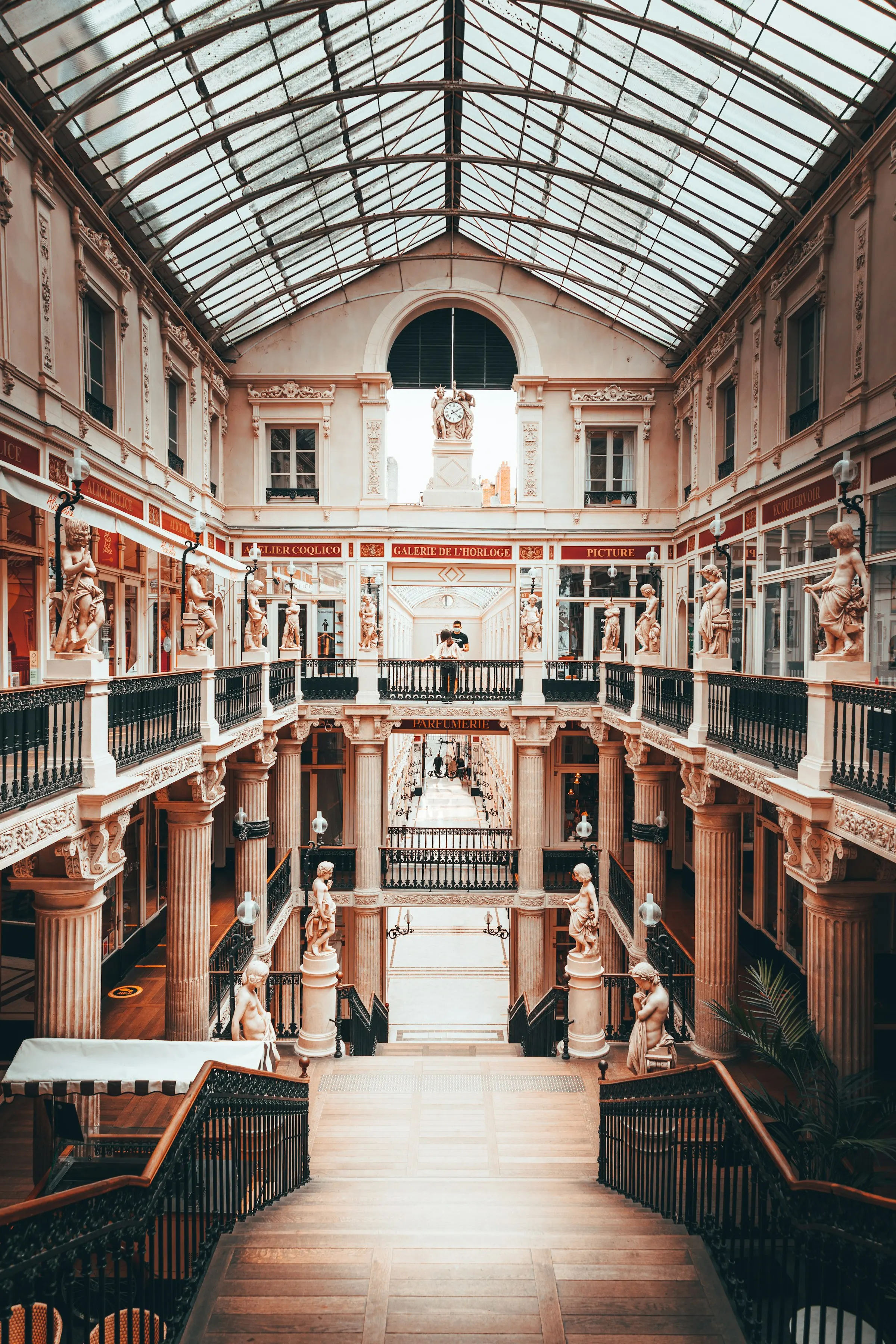 Inside the Passage Pommeraye, Nantes's ornate 19th-century glass-roofed shopping arcade, with tiered iron balustrades, classical statues and a grand wooden staircase descending between the softly lit levels.