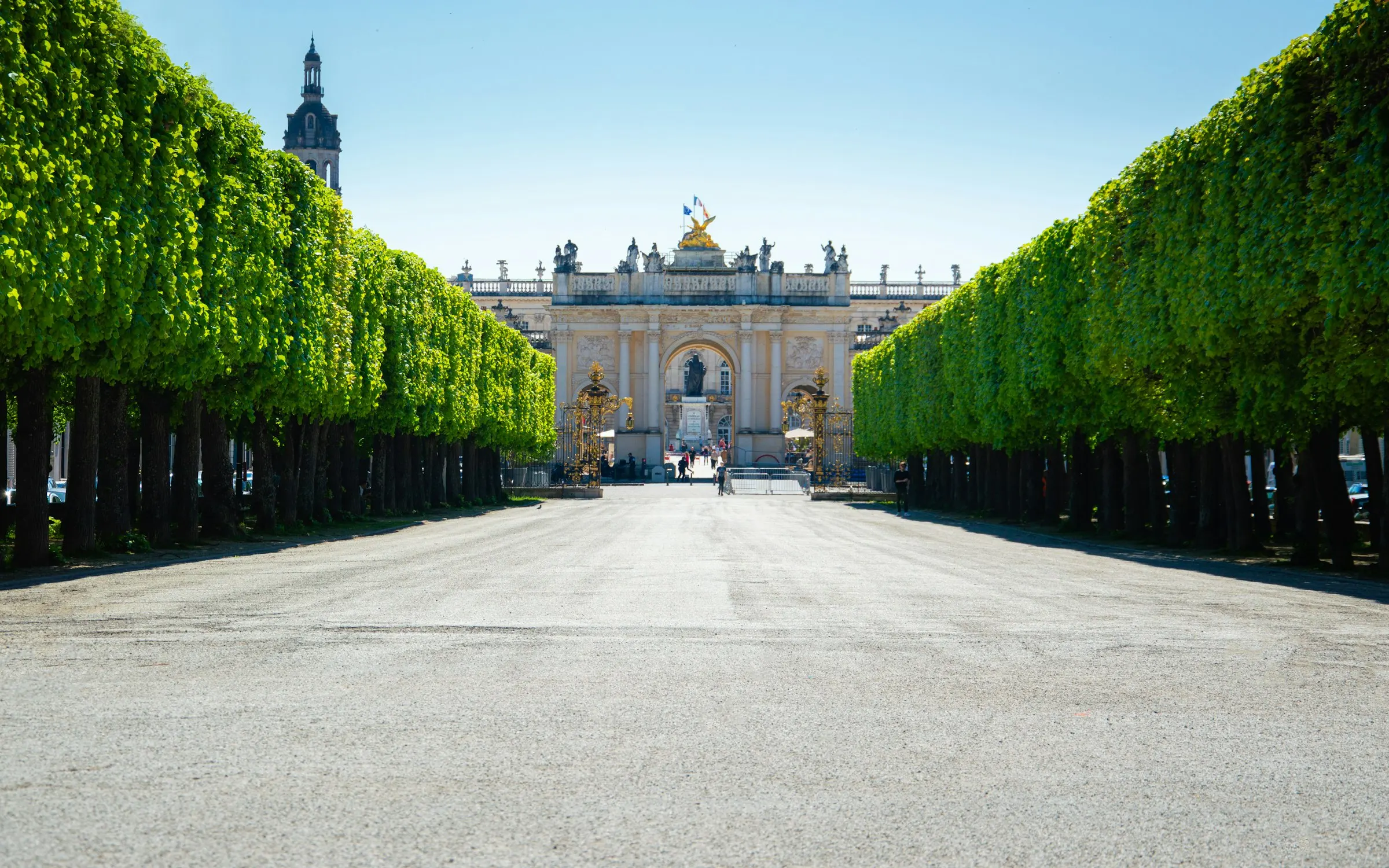 A pleached lime-tree allee in Nancy's Pepiniere park leading the eye to the triumphal Arc Here and gilded gates crowned by a golden winged statue, under a clear blue summer sky