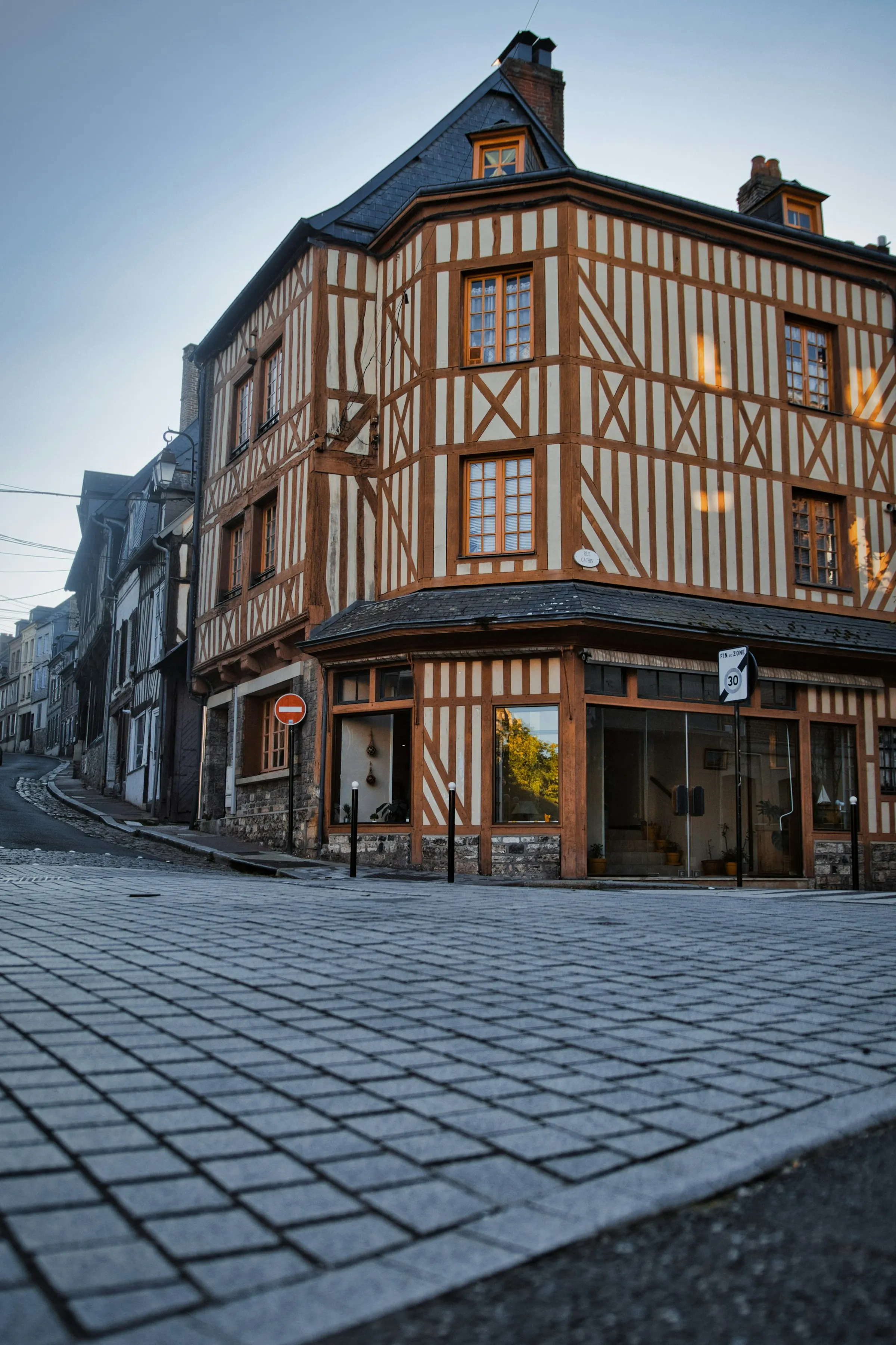 A handsome half-timbered corner building on a quiet cobbled street in Honfleur's old town, its timber framing glowing in early-morning golden light