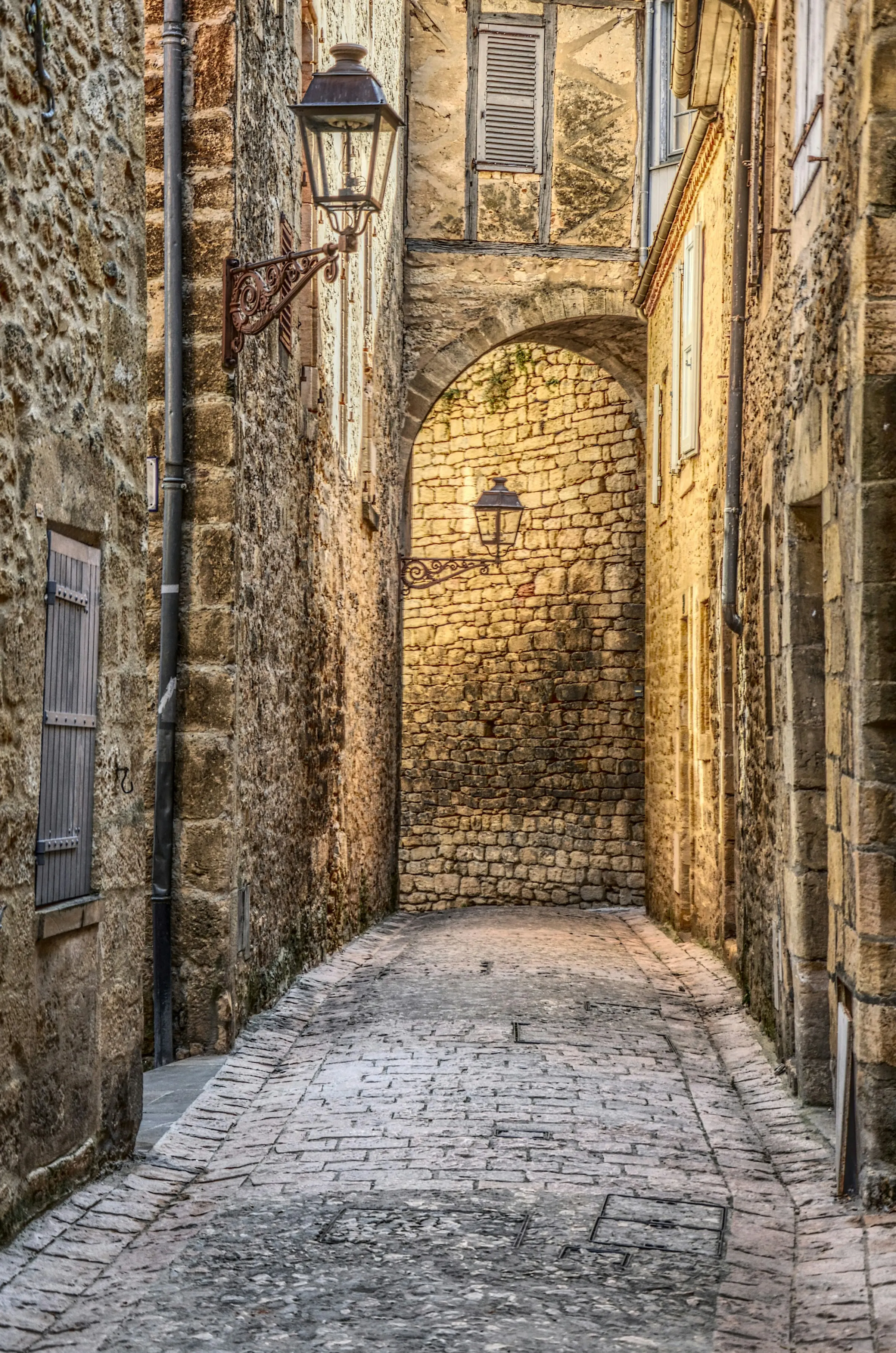 A quiet cobblestone lane in Sarlat framed by a golden-stone arched passageway, an old iron lantern against warm sunlit limestone walls