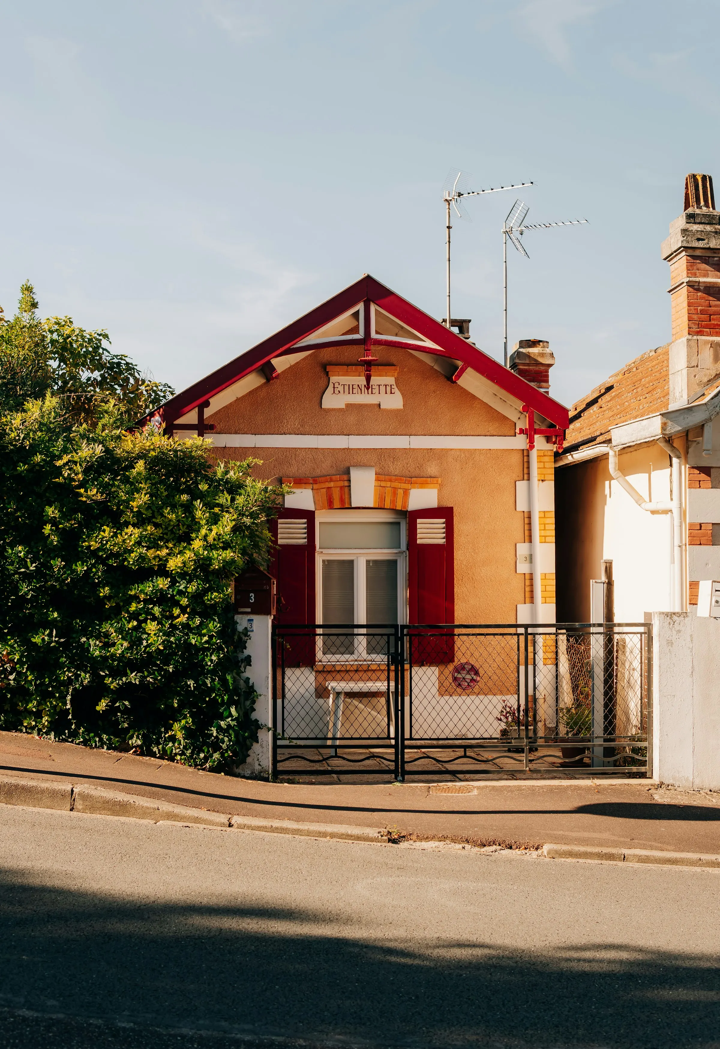 A small arcachonnaise seaside villa named 'Etiennette' with deep-red shutters and a carved wooden gable, glowing in warm golden evening light behind a low iron gate.