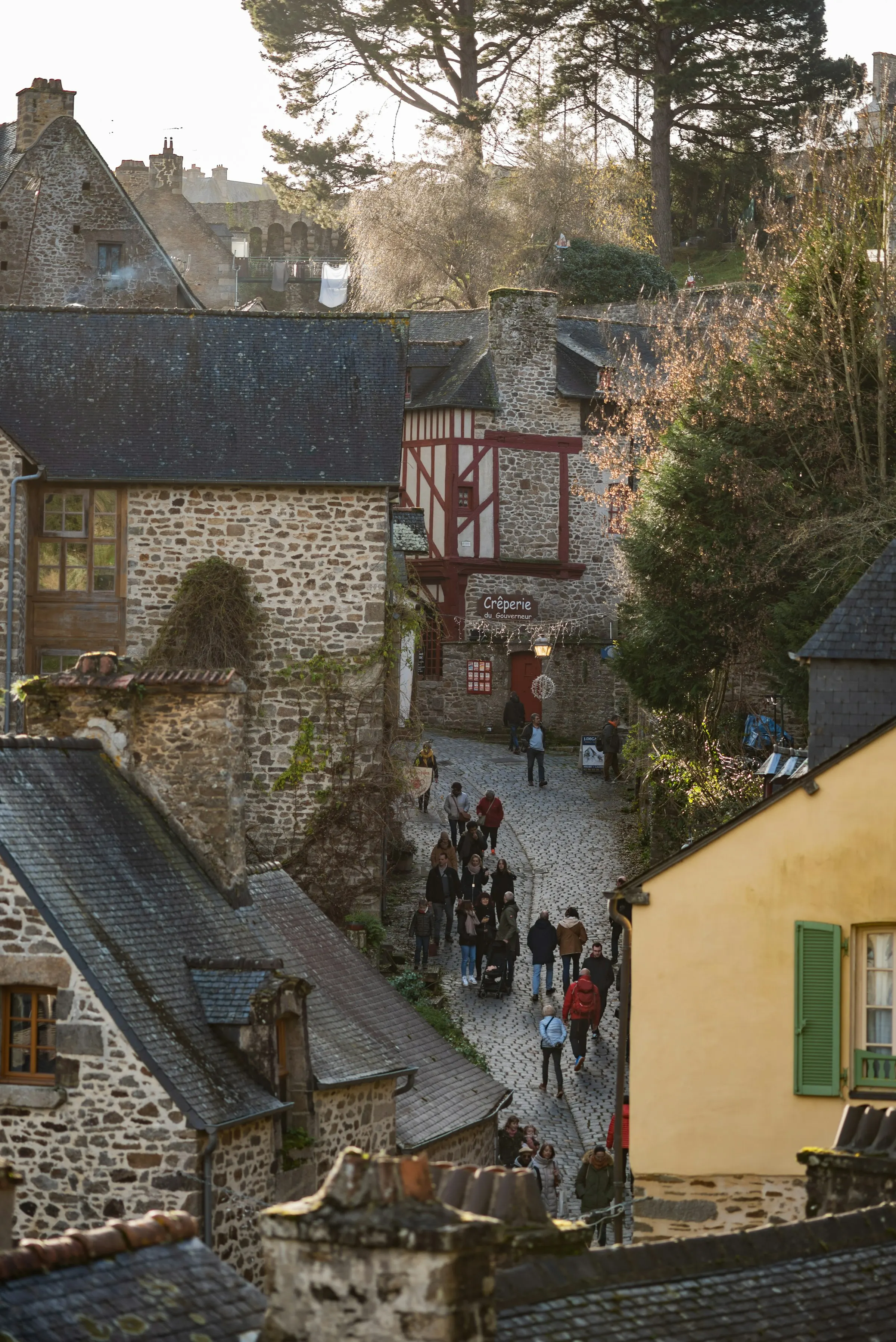 The steep cobbled Rue du Jerzual descending between half-timbered stone houses, a red-fronted Creperie du Gouverneur, backlit by low winter sun with walkers heading down