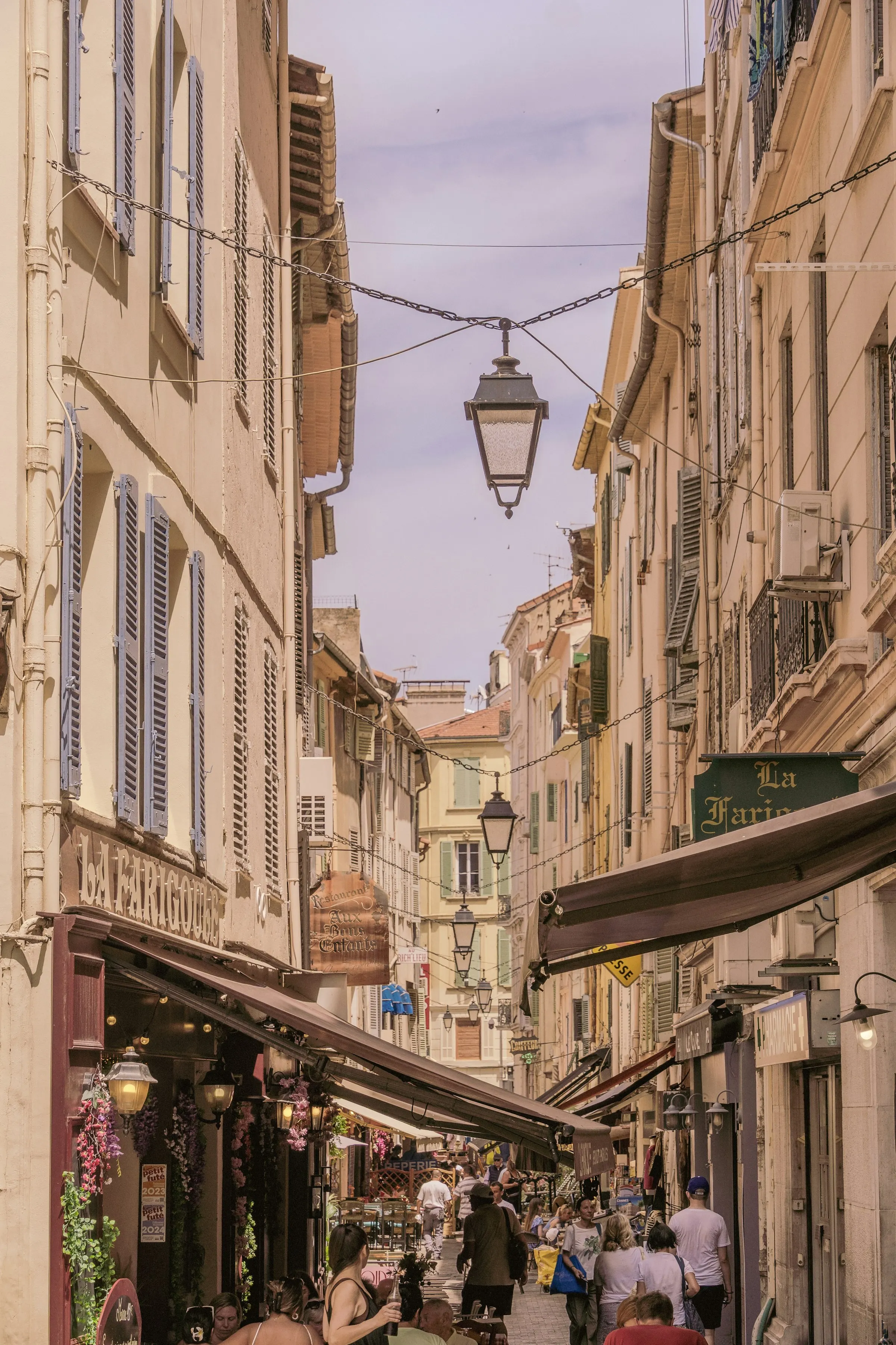 A lively pedestrian lane in the old town of Cannes, awnings and wrought-iron lanterns strung between tall shuttered houses, cafes and food shops with diners at outdoor tables in warm afternoon light.