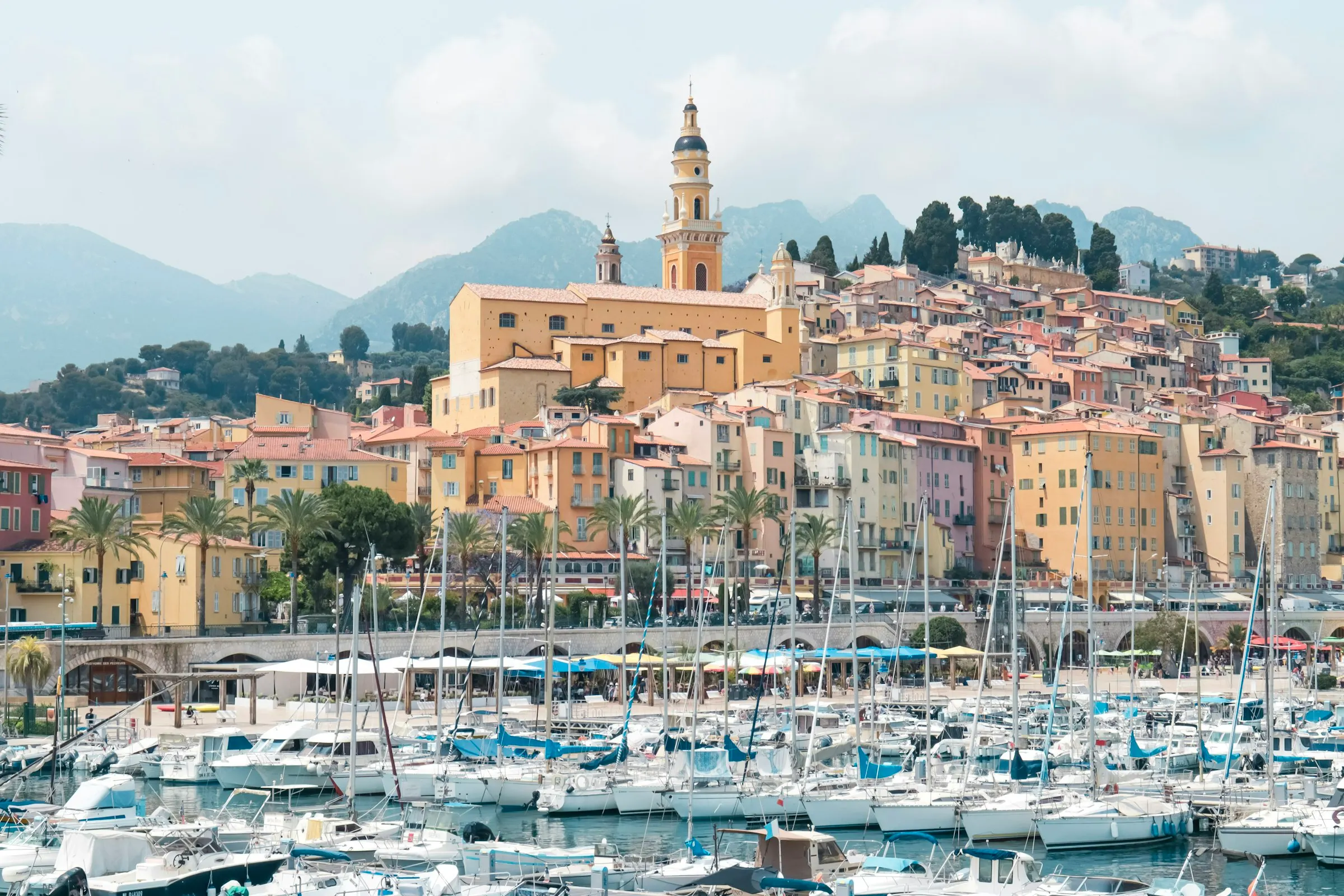 Menton's old port packed with white sailing yachts, the colorful hillside old town and the ochre Saint-Michel bell tower rising behind beneath misty coastal mountains.
