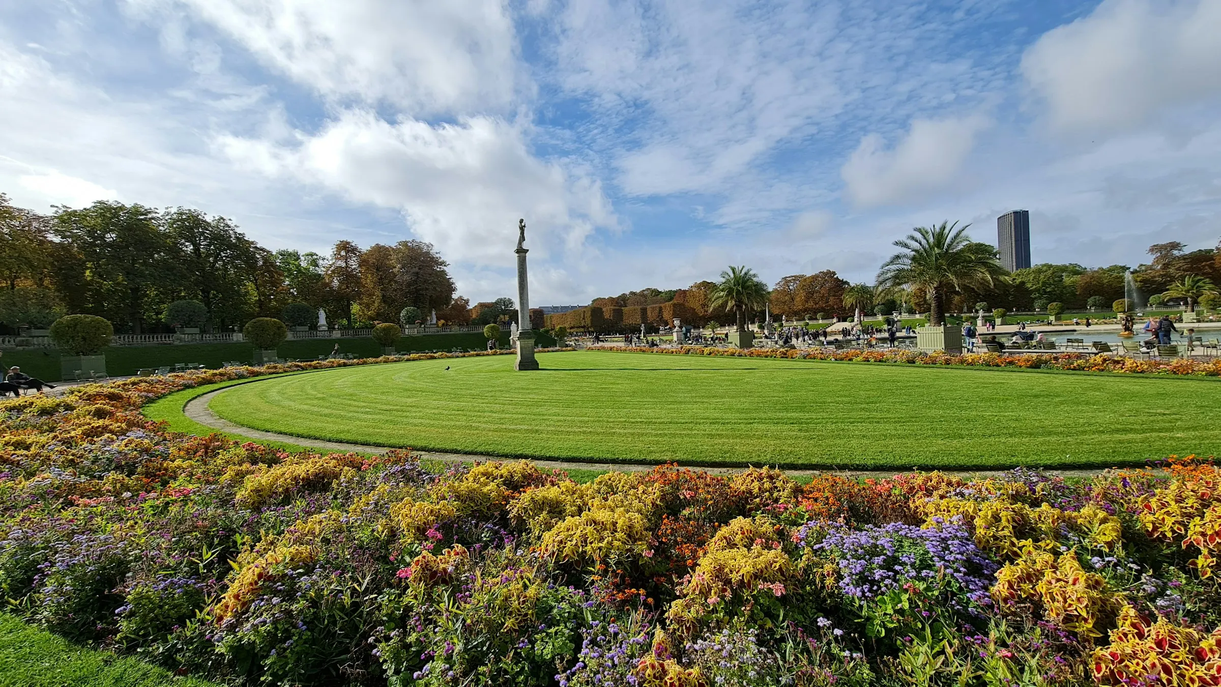 The Jardin du Luxembourg on a bright day — a circular lawn ringed with autumn flowerbeds and palm trees, with Parisians strolling and sitting out