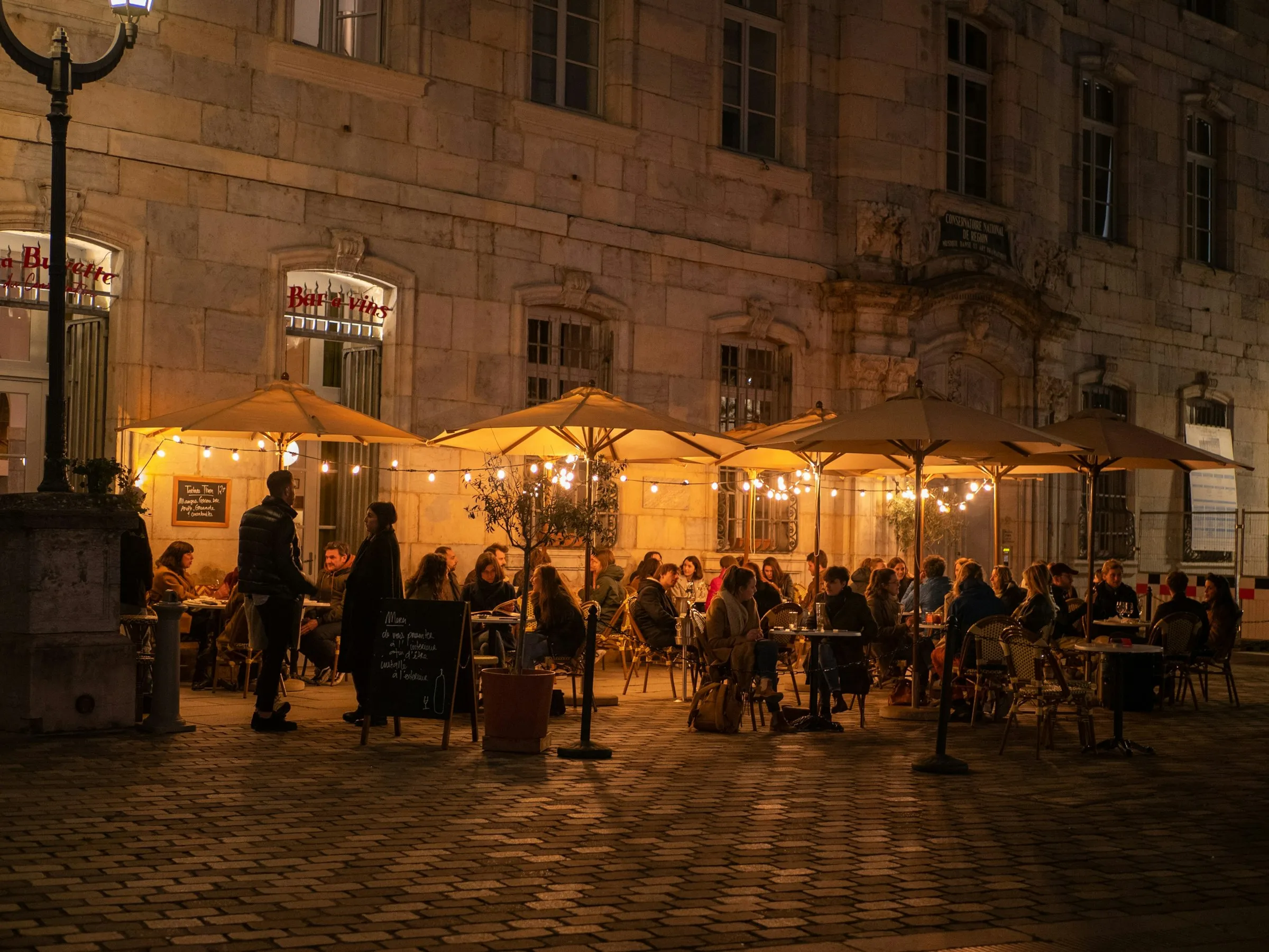 Evening café terrace on a cobbled Besançon square, cream canvas umbrellas strung with warm fairy lights and diners at bistro tables against the floodlit honey-stone façade of the old Conservatoire.