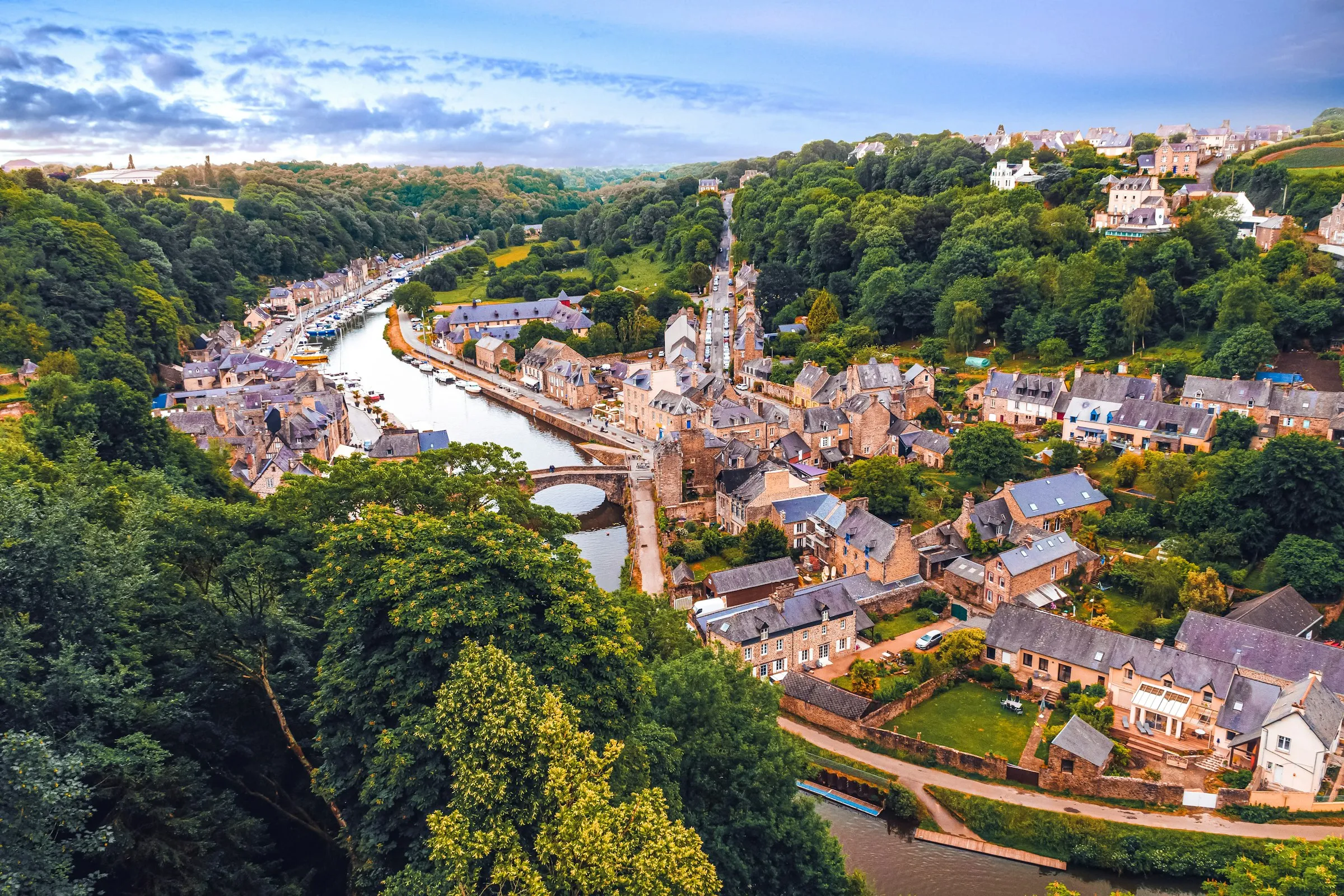 Golden-hour aerial view of Dinan's medieval stone town nestled in the wooded Rance river valley, with the old arched bridge and the port lined with boats below