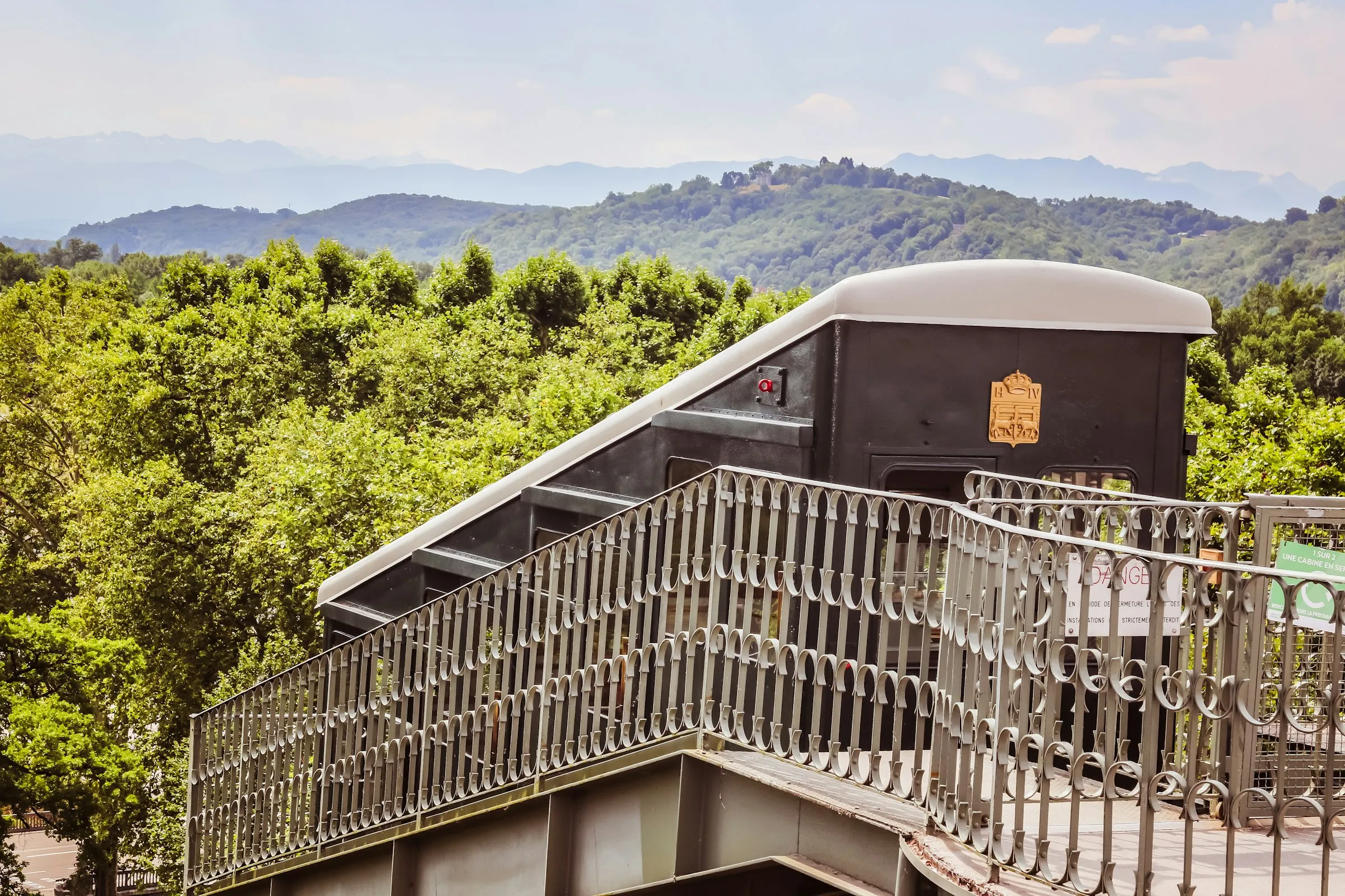 The historic black-and-cream cabin of the Funiculaire de Pau, marked with the gilded Henri IV coat of arms of Béarn, cresting its track above the treetops with the hazy blue ridges of the Pyrenees foothills rolling into the distance under a bright summer sky