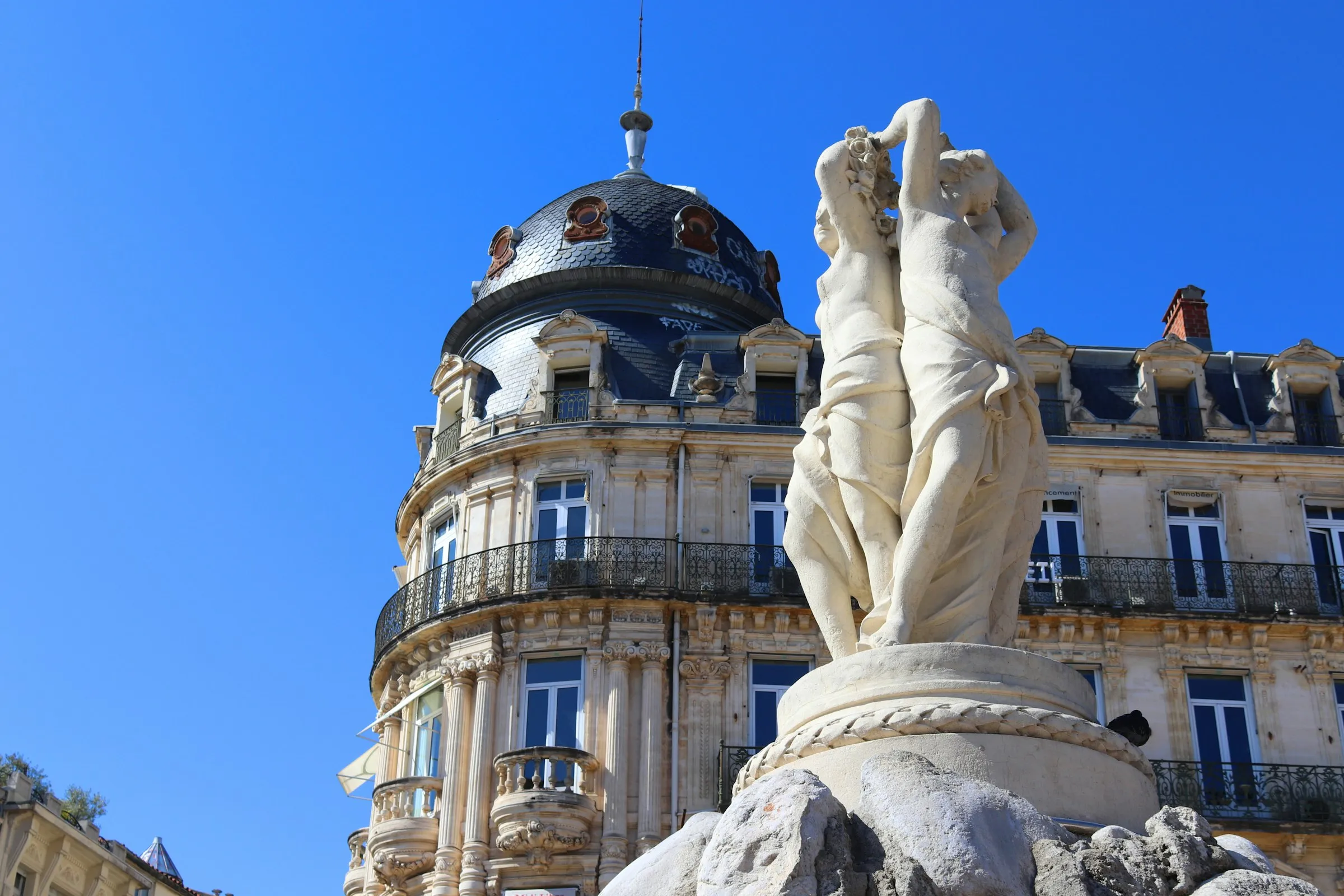 The Three Graces fountain on Place de la Comedie in Montpellier, the city iconic central square, below the domed Gaumont-Comedie building under a deep blue sky