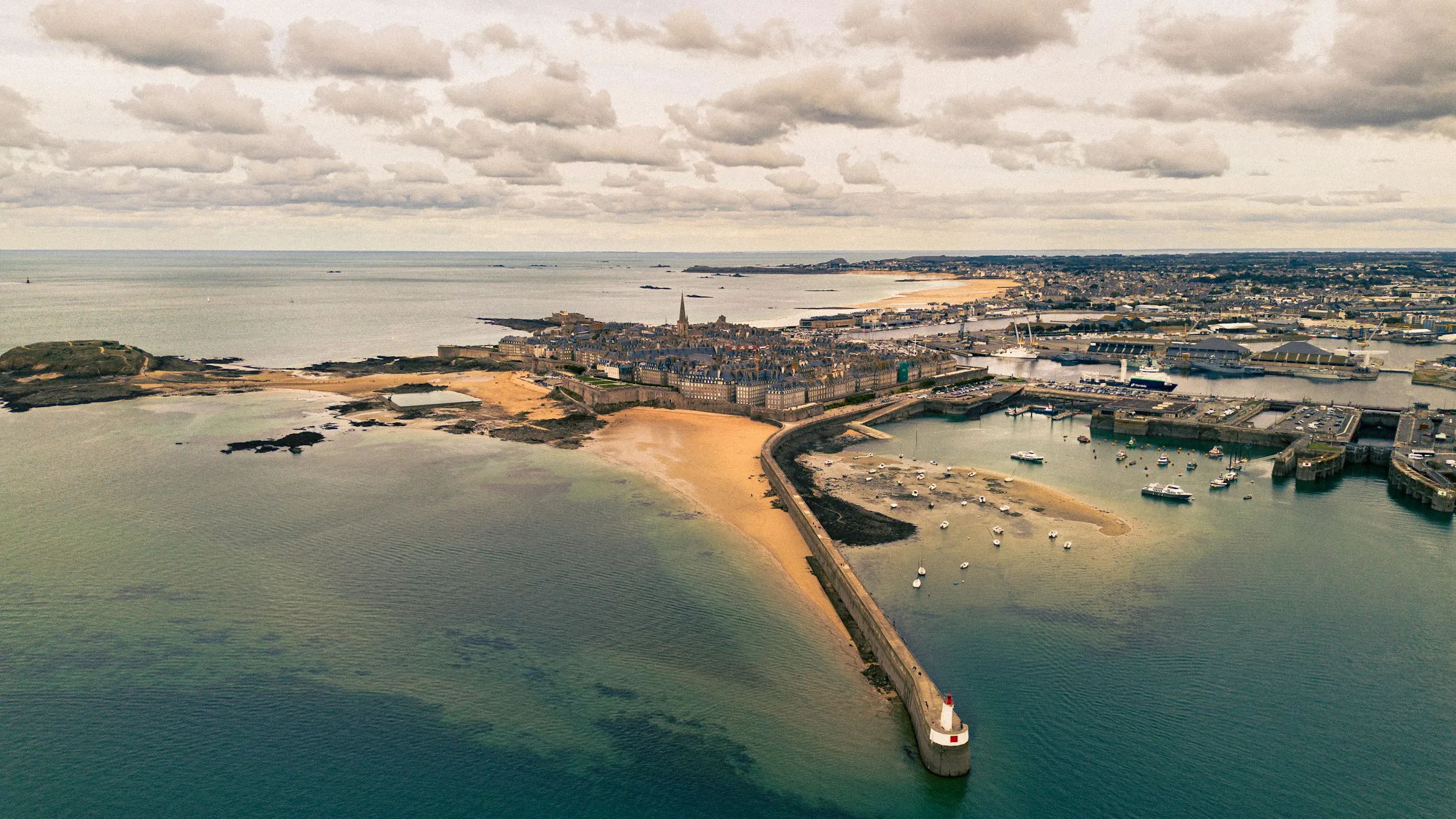 Aerial view of Saint-Malo's walled granite old town jutting into the sea, encircled by ramparts, with sandy tidal beaches, the yacht harbour and the Mole des Noires lighthouse jetty under a cloud-scattered sky.