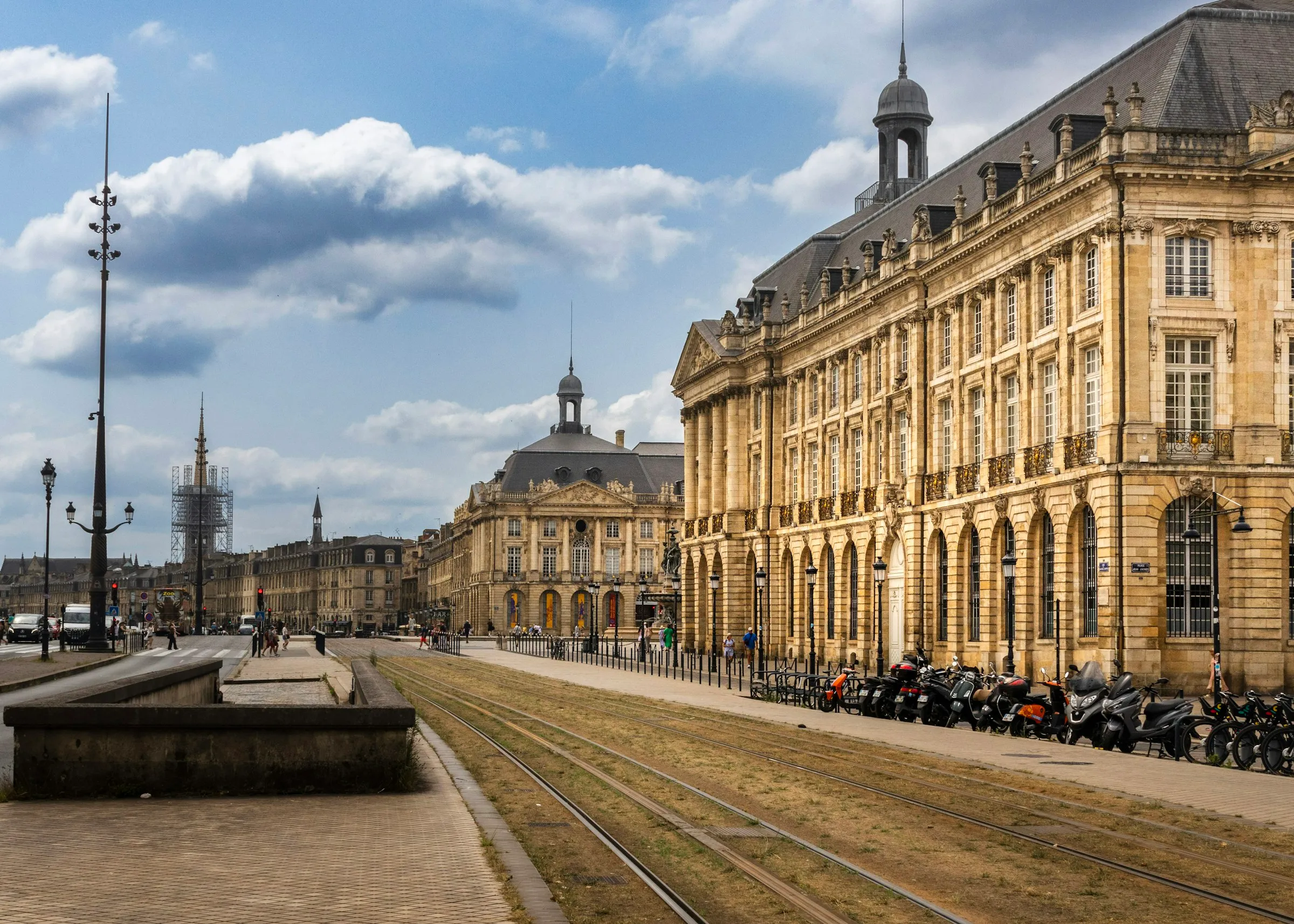 The grand 18th-century Palais de la Bourse along Bordeaux's quays in warm afternoon light, a sweep of pale-limestone facade with arched windows and a domed corner pavilion, tram tracks running past under a blue sky with drifting clouds.