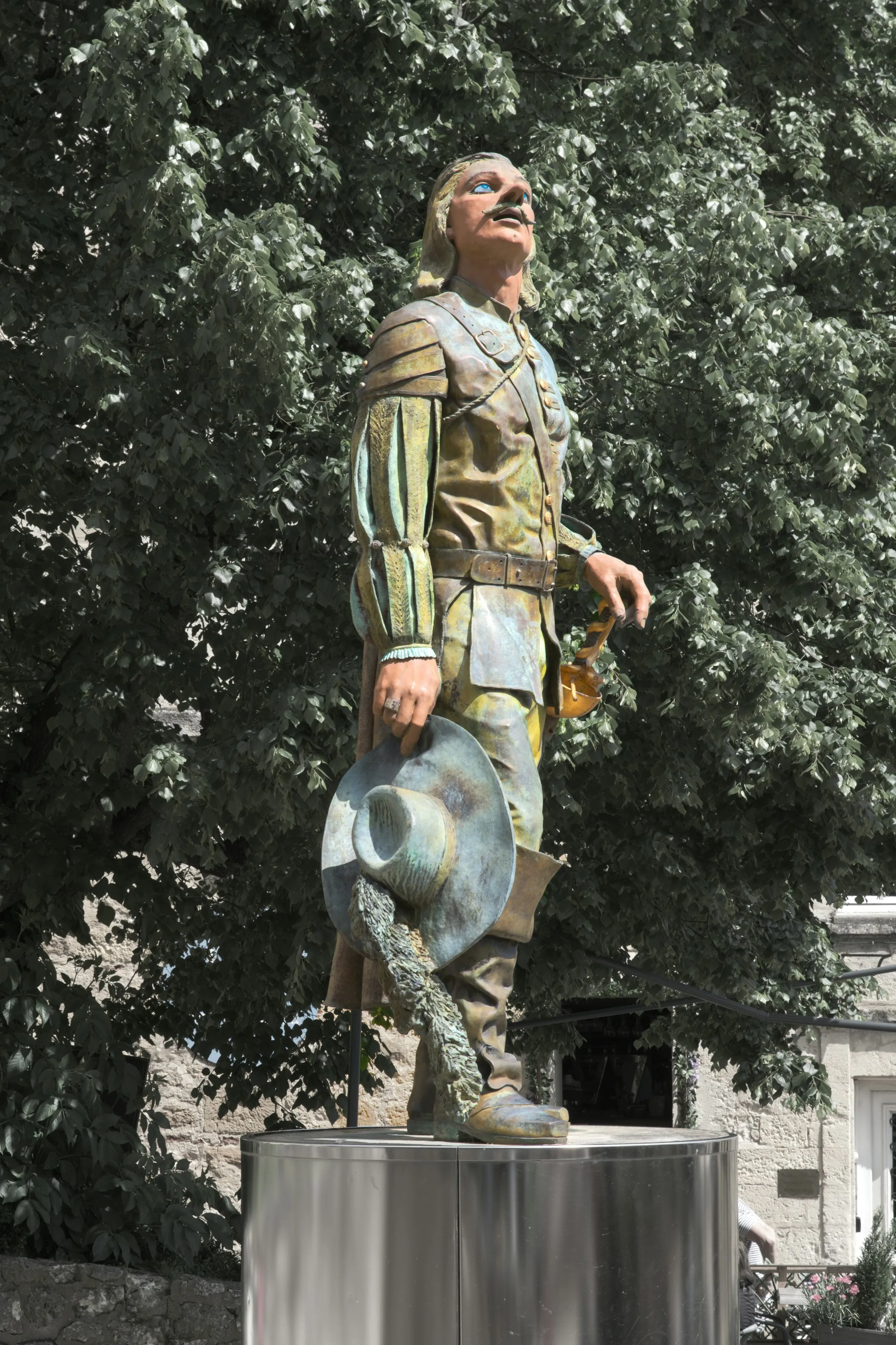 The bronze Cyrano de Bergerac statue in the old town, plumed hat in hand and gaze raised, framed by bright summer foliage — the emblem of the town