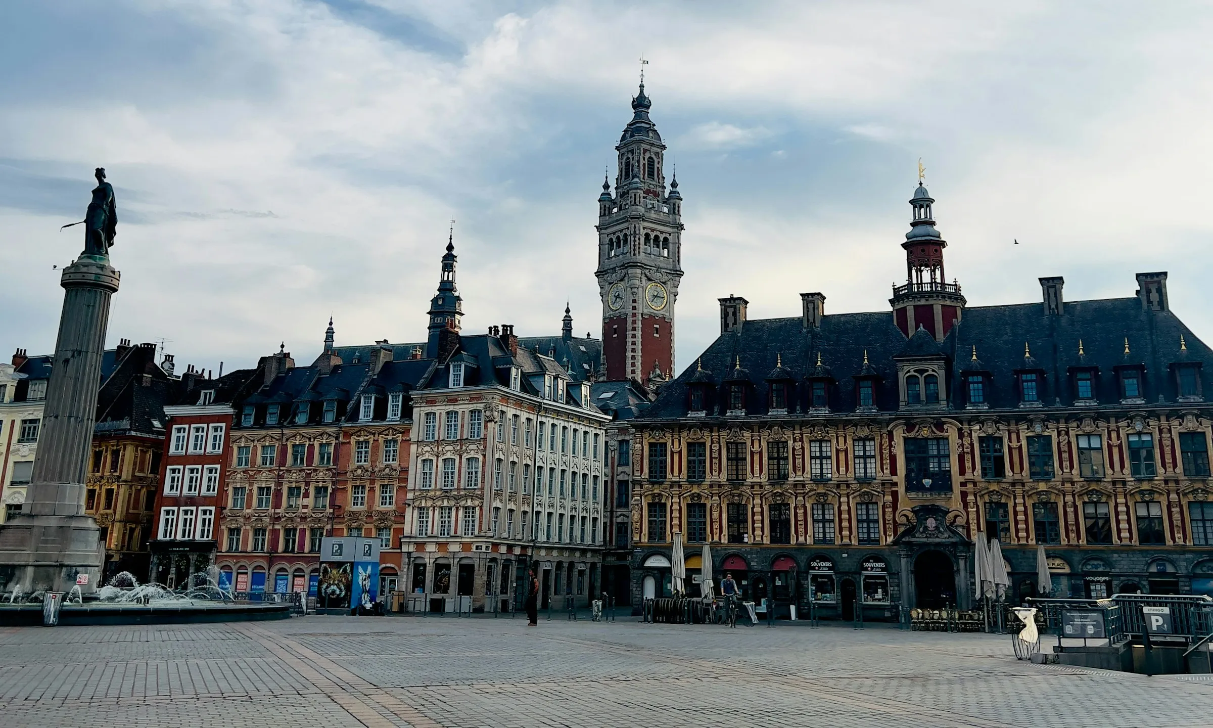 Lille's Grand Place in soft daylight, the tall Chamber of Commerce belfry rising behind ornate Flemish-Baroque gabled facades, the columned Vieille Bourse, and the Deesse column with its fountain on the wide cobbled square.