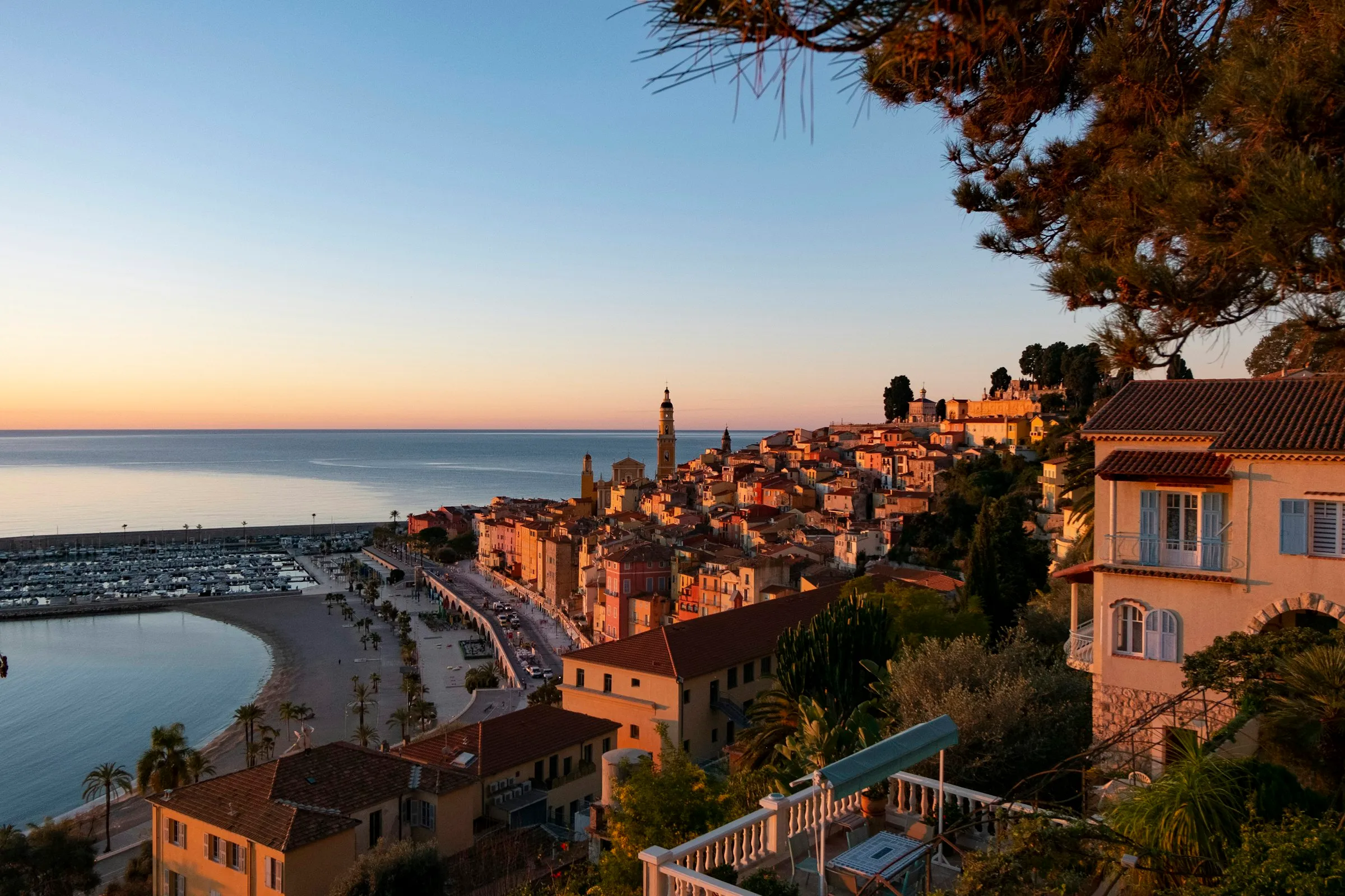 Golden-hour view over Menton's old town from a hillside villa terrace, the ochre Saint-Michel bell tower rising above stacked pastel houses, the curved beach and yacht marina running along a calm sea.