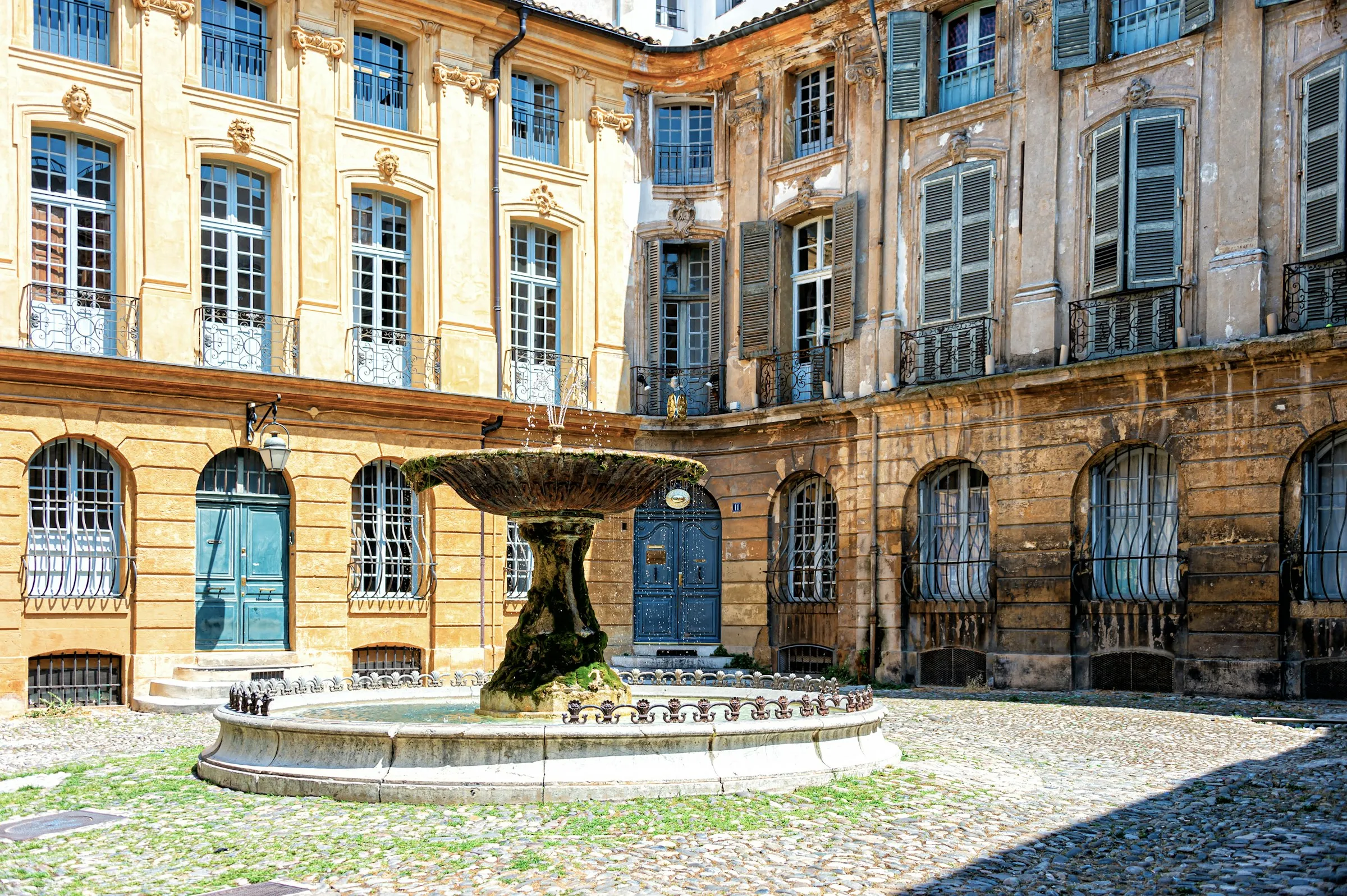 Sunlit Place d'Albertas in Aix-en-Provence: a moss-covered stone fountain on a cobbled square ringed by ornate 18th-century golden-stone facades with pastel shutters and wrought-iron balconies.