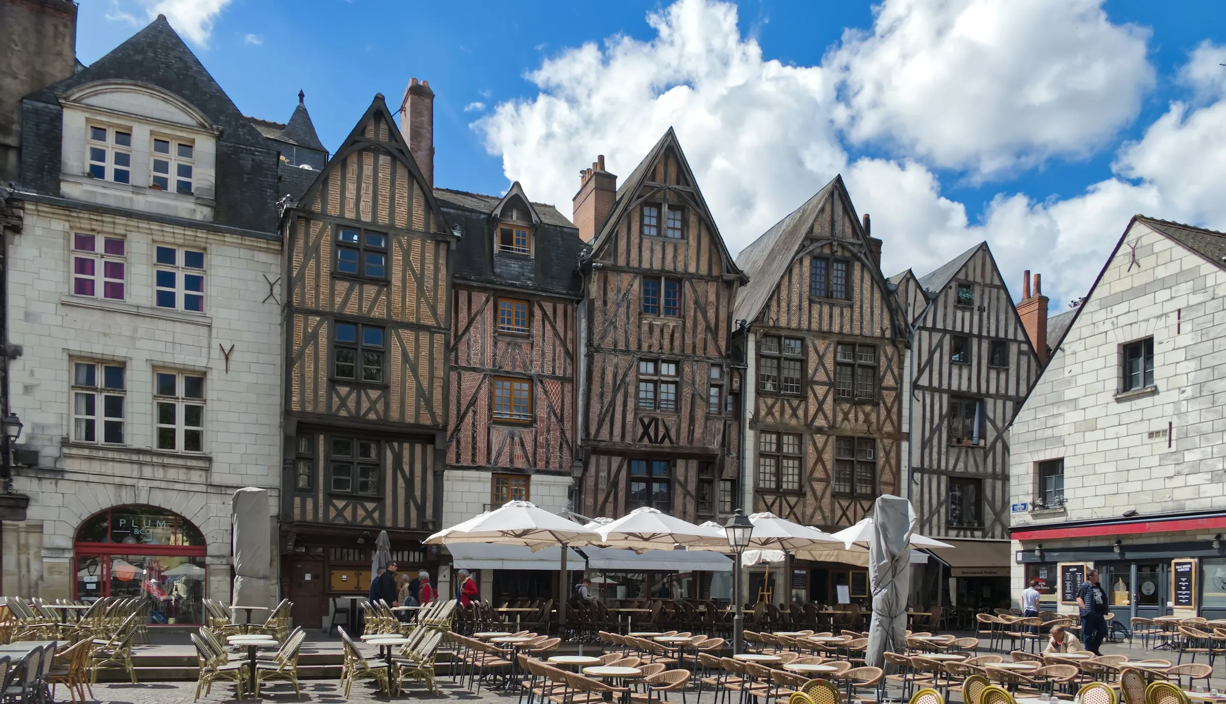Sunlit medieval half-timbered houses lining Place Plumereau in the old town of Tours, with café tables and white parasols set out across the square beneath a bright blue sky full of billowing white clouds.