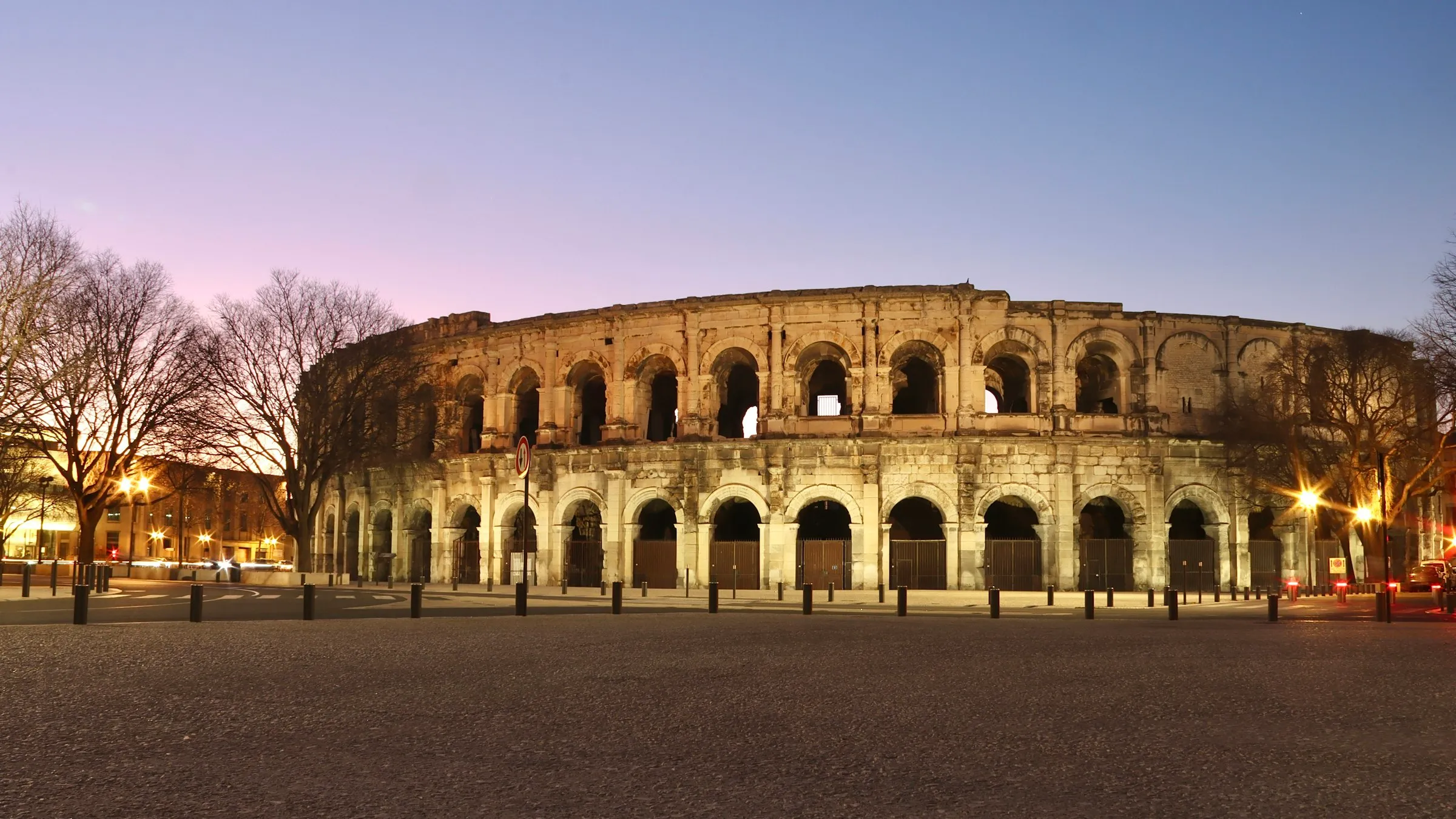 The Roman amphitheatre of Nimes (Arenes de Nimes) at dusk, its two tiers of stone arcades glowing warm gold against a violet-to-blue twilight sky, ringed by bare winter plane trees, streetlights, and an empty paved plaza.