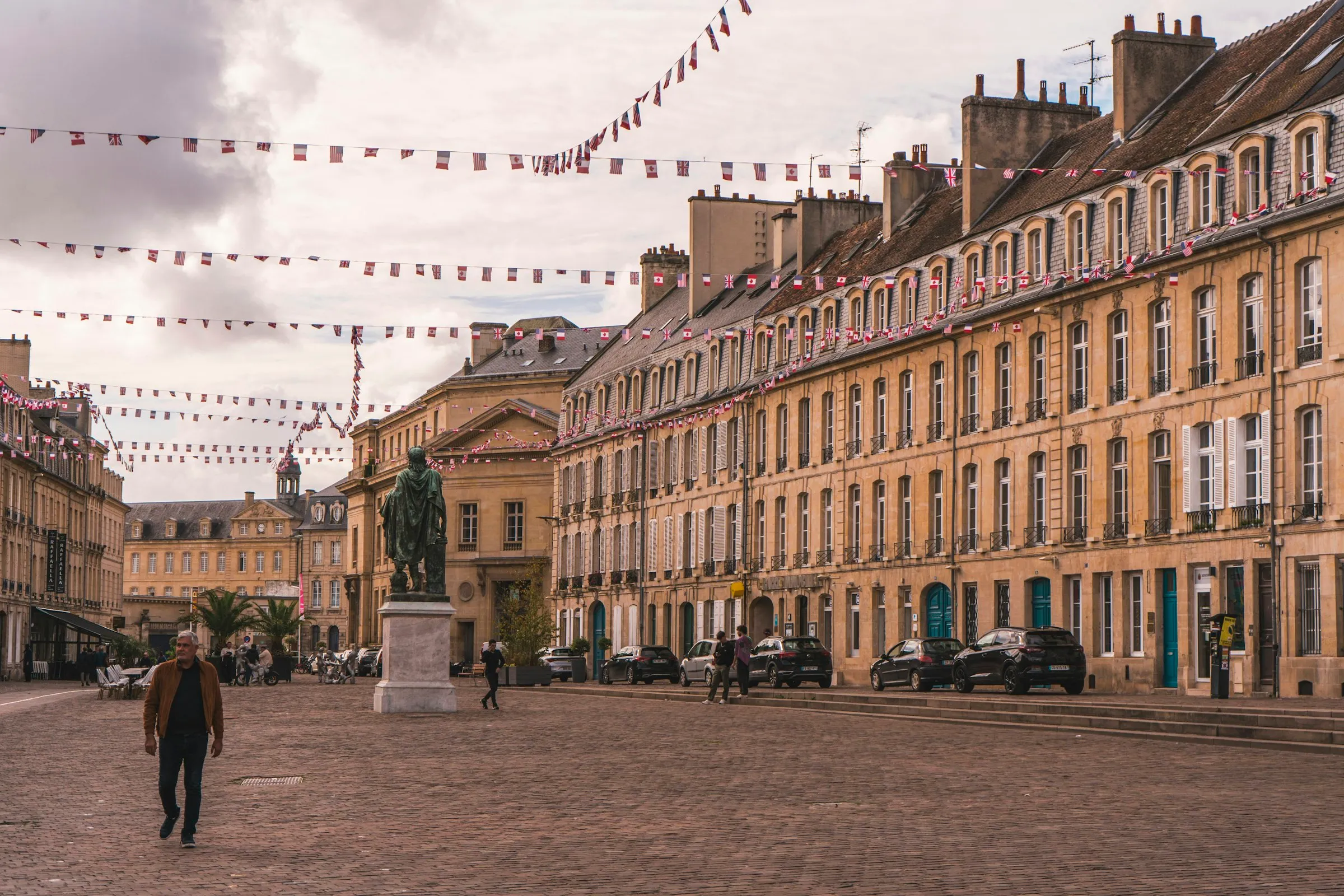 The Place Saint-Sauveur in the old town of Caen, Normandy — an elegant cobbled square lined with honey-stone 18th-century townhouses and strung with French, British, American and Canadian bunting, with the bronze statue of Louis XIV at its centre