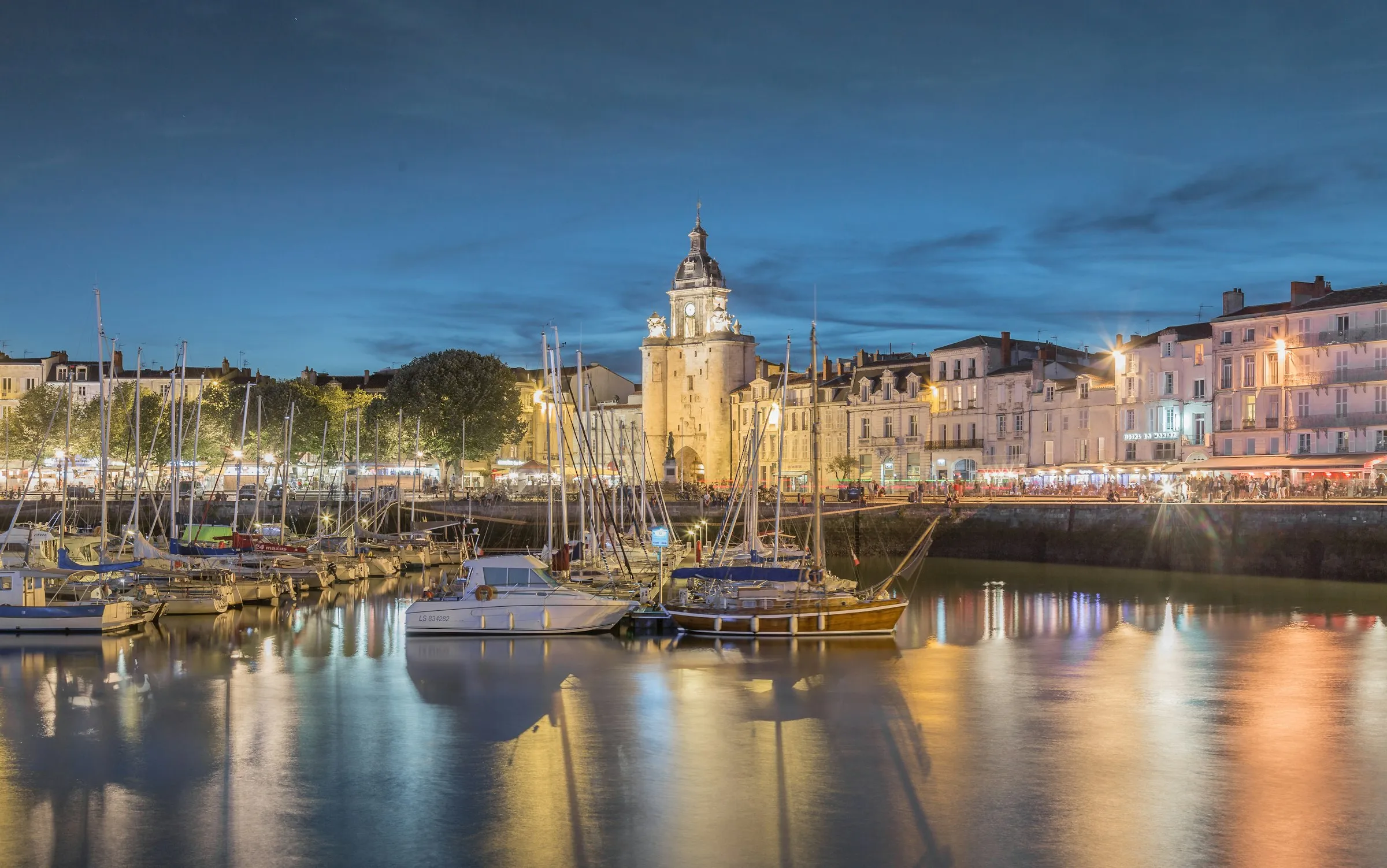 La Rochelle's Vieux-Port at blue hour, the floodlit Grosse Horloge clock-tower gate and lamplit quayside restaurants mirrored in the still water among rows of moored yachts.